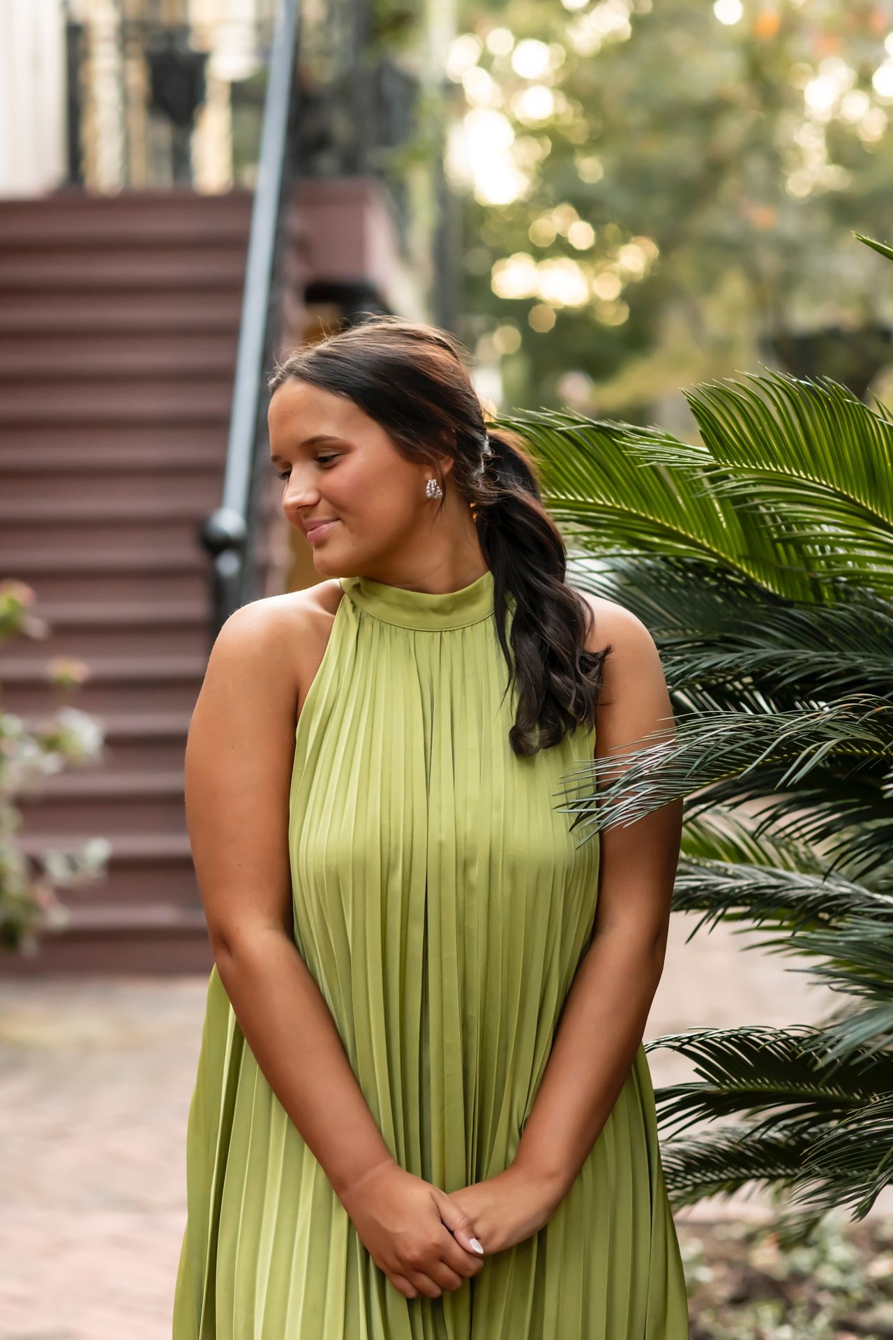 Woman in lime green pleated dress poses outdoors near palm plants in natural lighting.