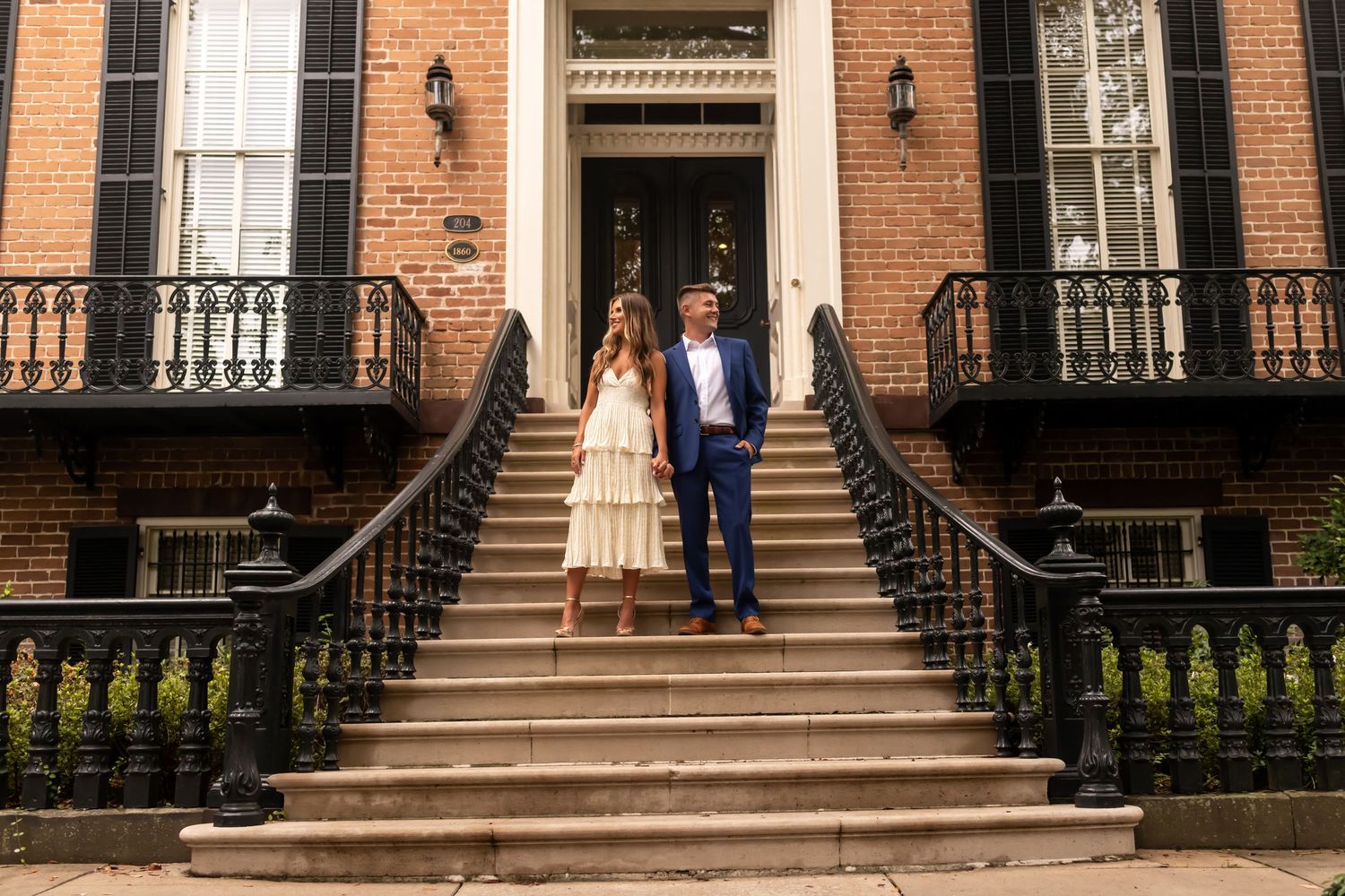 A couple stands together on historic brownstone steps with ornate black railings and brick facade in an elegant urban setting.