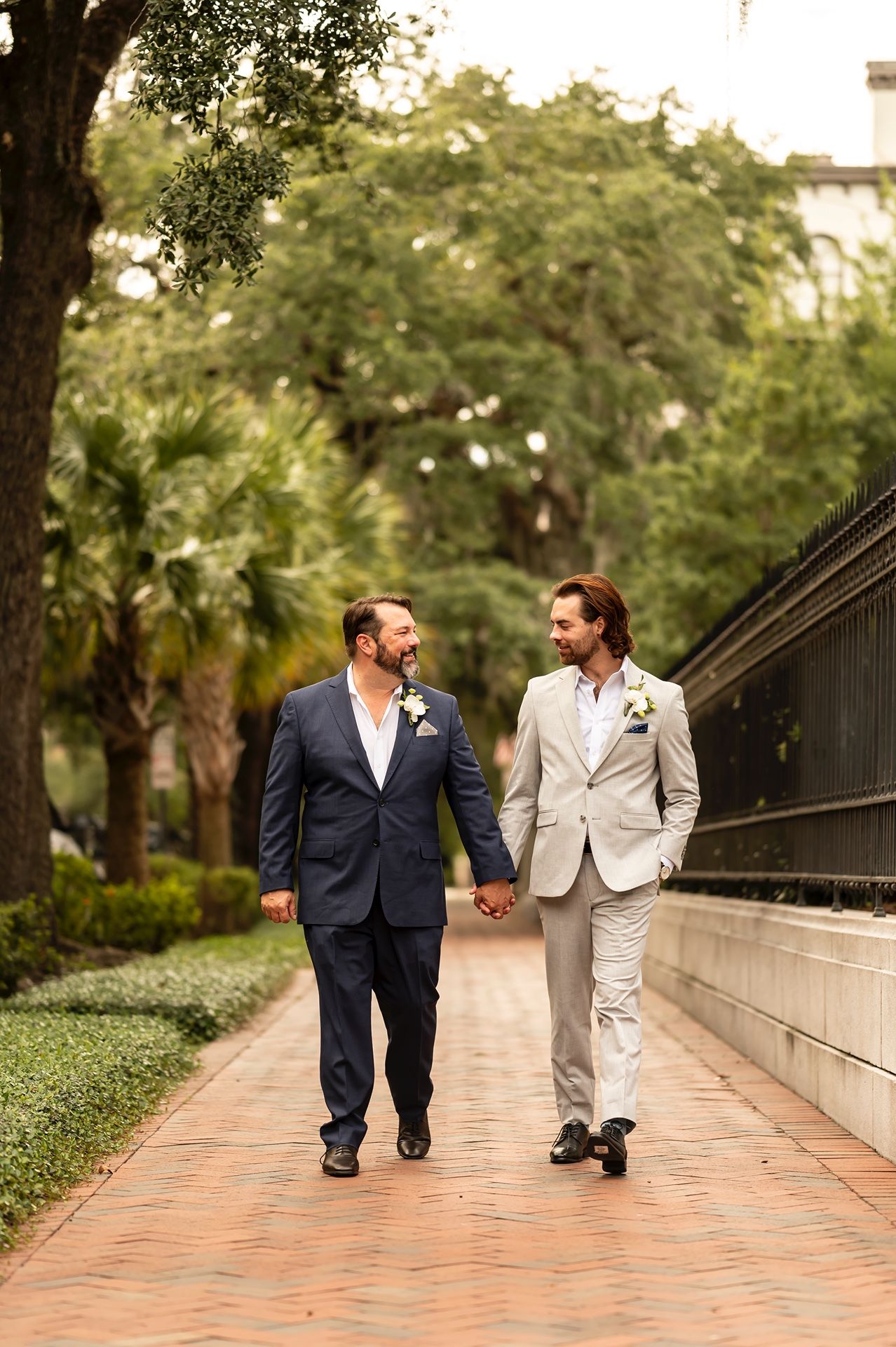 Two people in suits stroll together along a palm-lined brick walkway while sharing a tender moment.