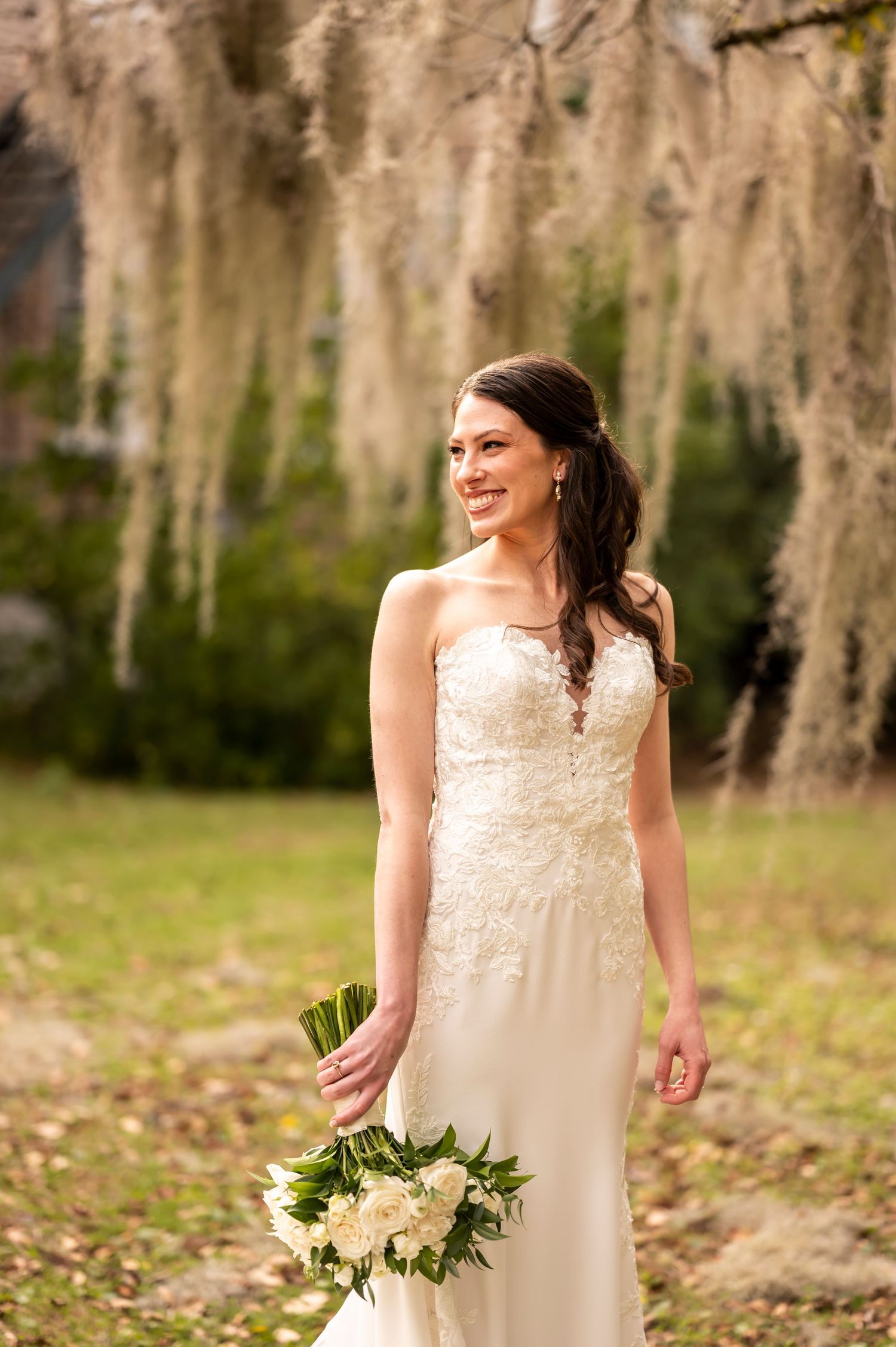 A bride in a strapless lace gown smiles while holding a white floral bouquet against a backdrop of Spanish moss.