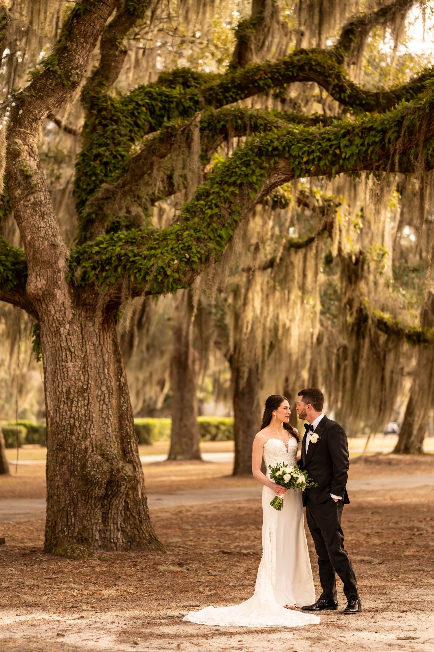 A couple shares an intimate moment under majestic oak trees draped with Spanish moss in a Southern garden setting.