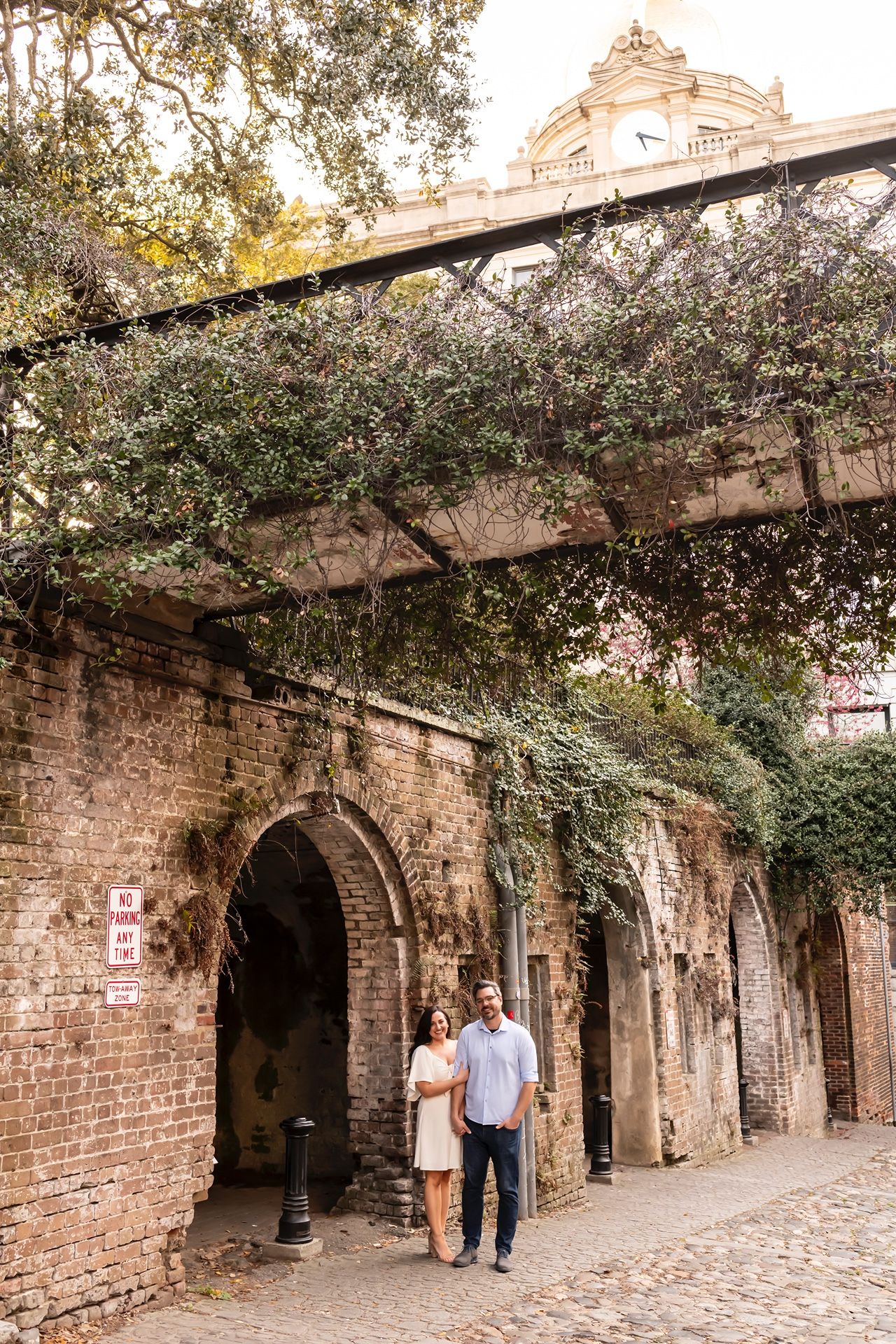 Romantic couple strolling under brick archways and tree-lined cobblestone path in historic Charleston district.