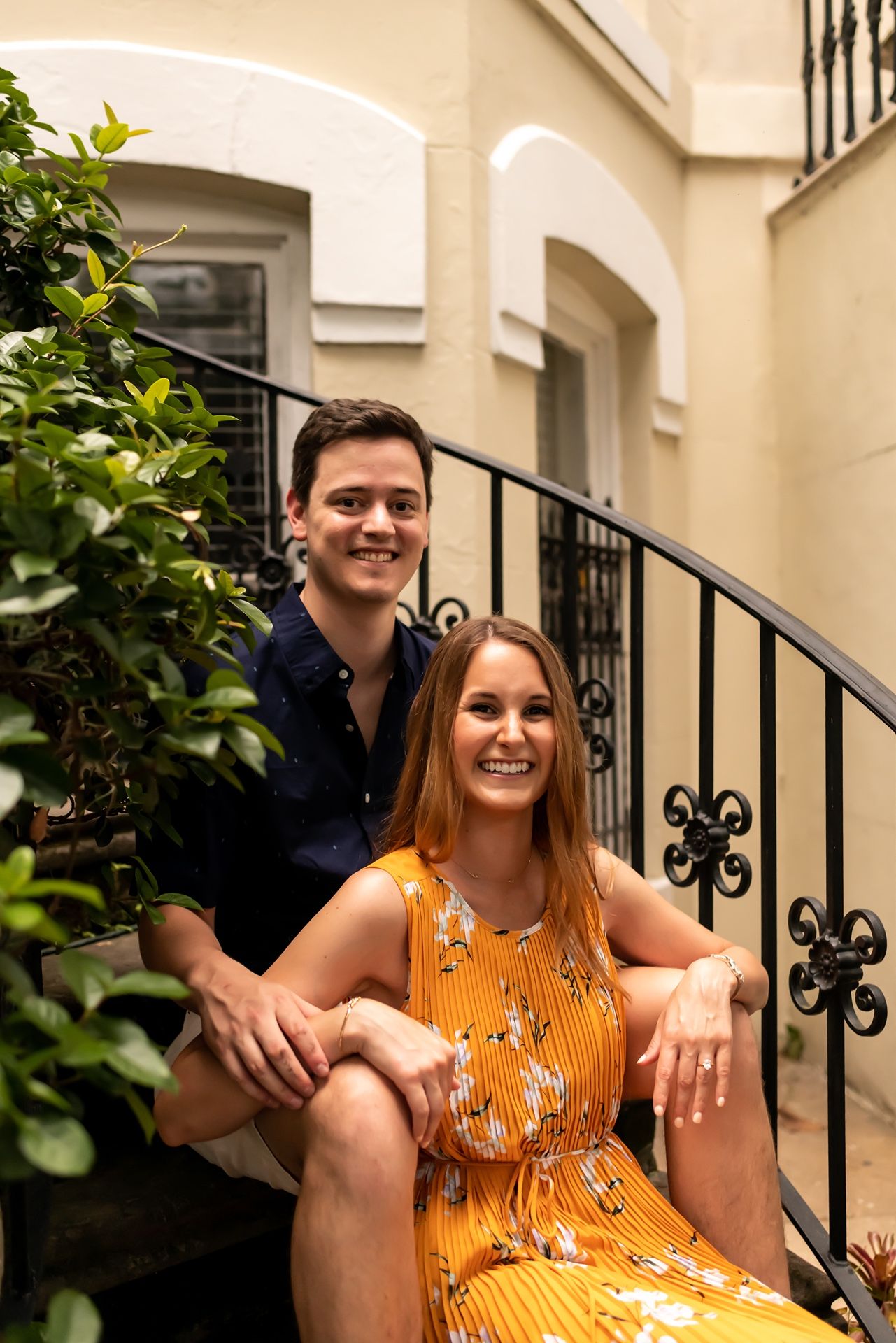 A couple sits together on outdoor stairs with decorative iron railings, near a beige stucco wall and green foliage.