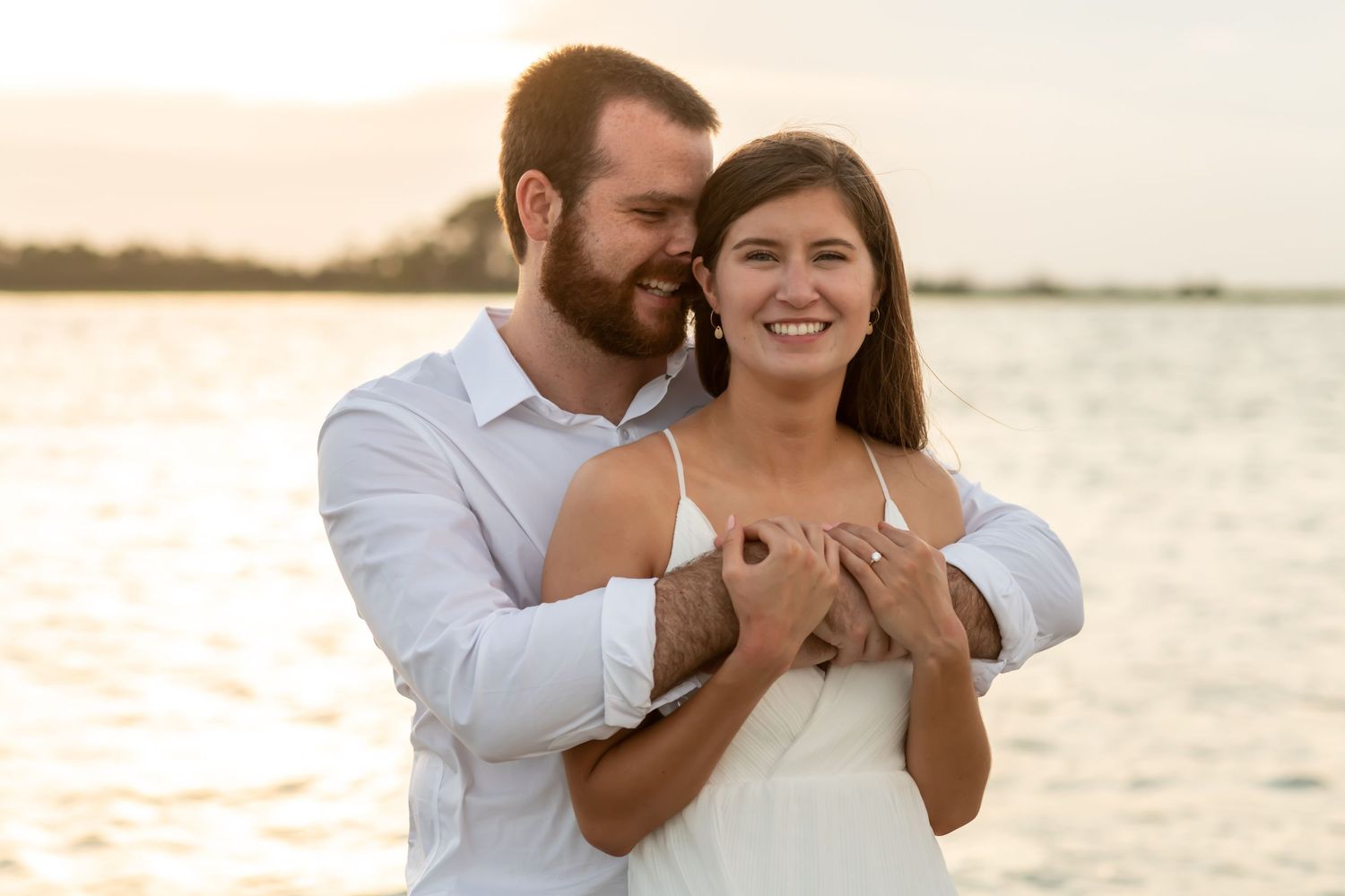 A happy couple embraces on a beach at sunset with water and golden light in the background.