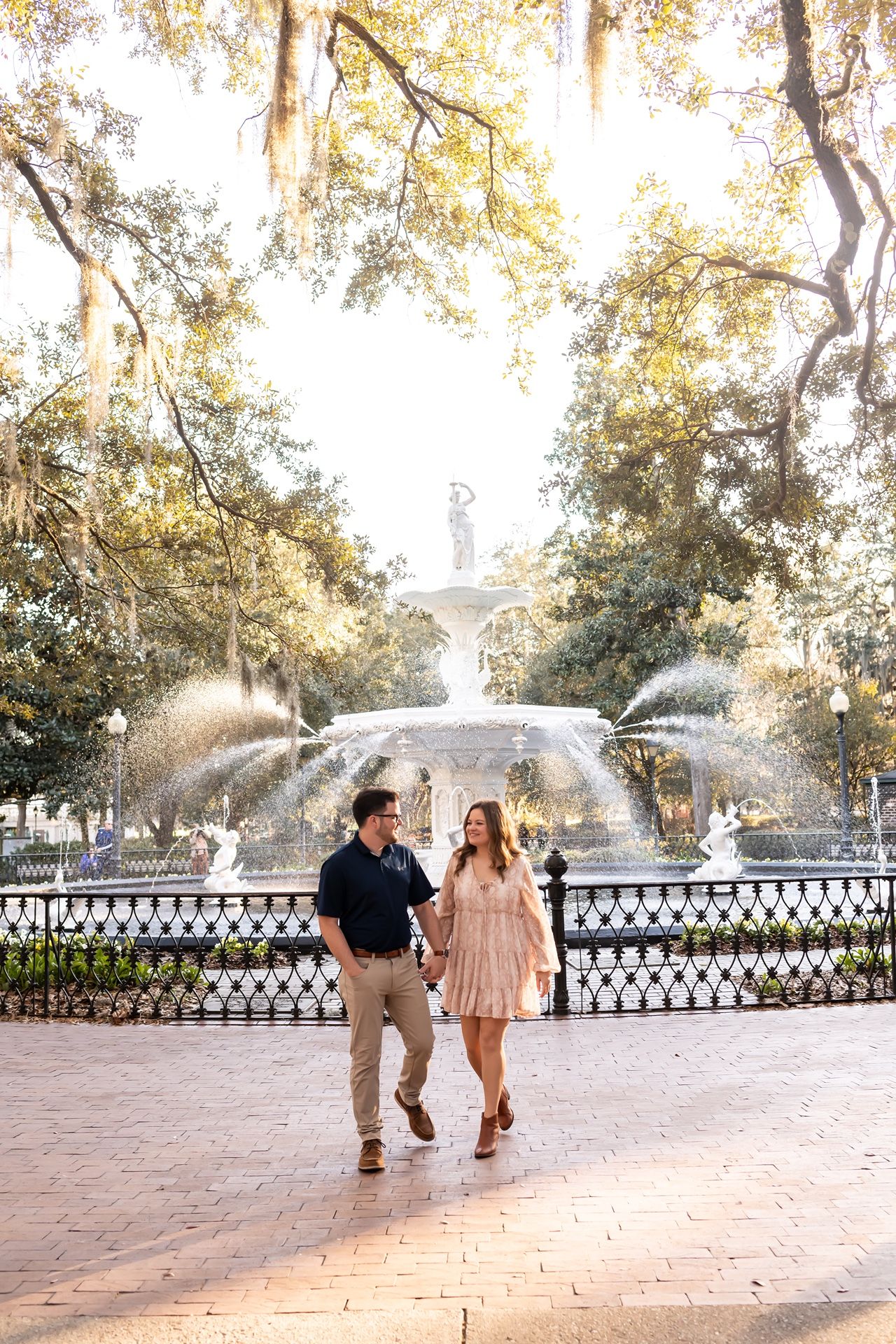 Couple strolls along brick pathway near ornate fountain in a lush park with Spanish moss draped trees and golden sunlight.
