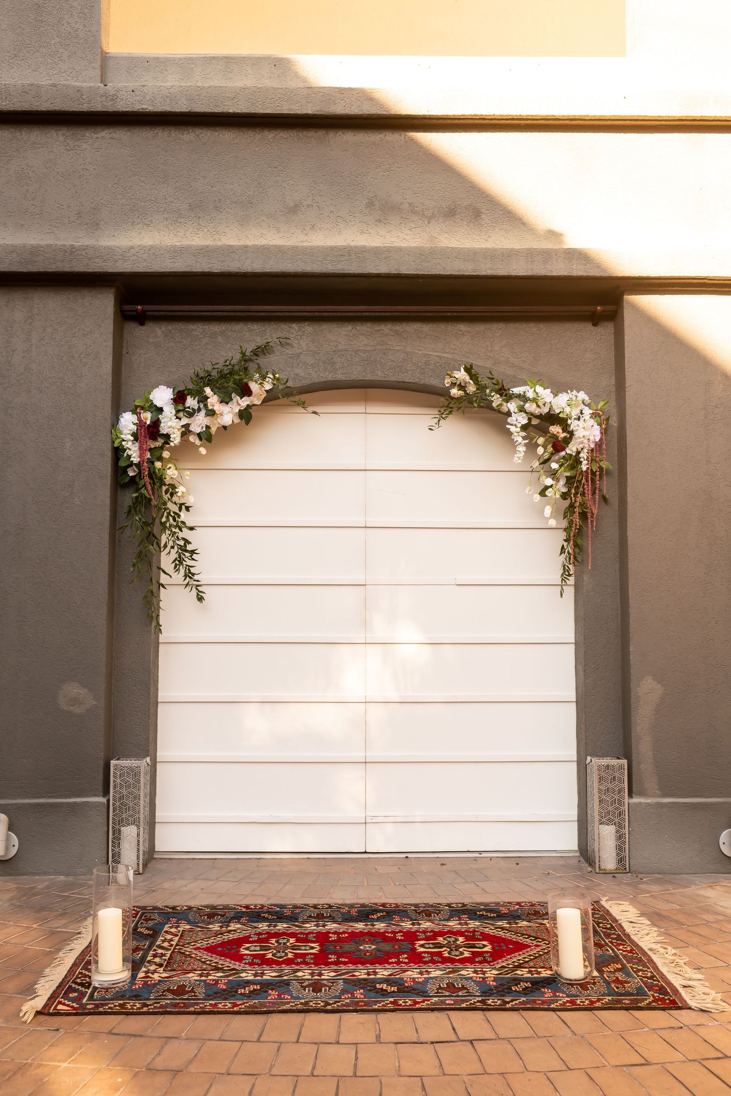 A white door decorated with floral garlands and candle pillars sits above a red patterned area rug.