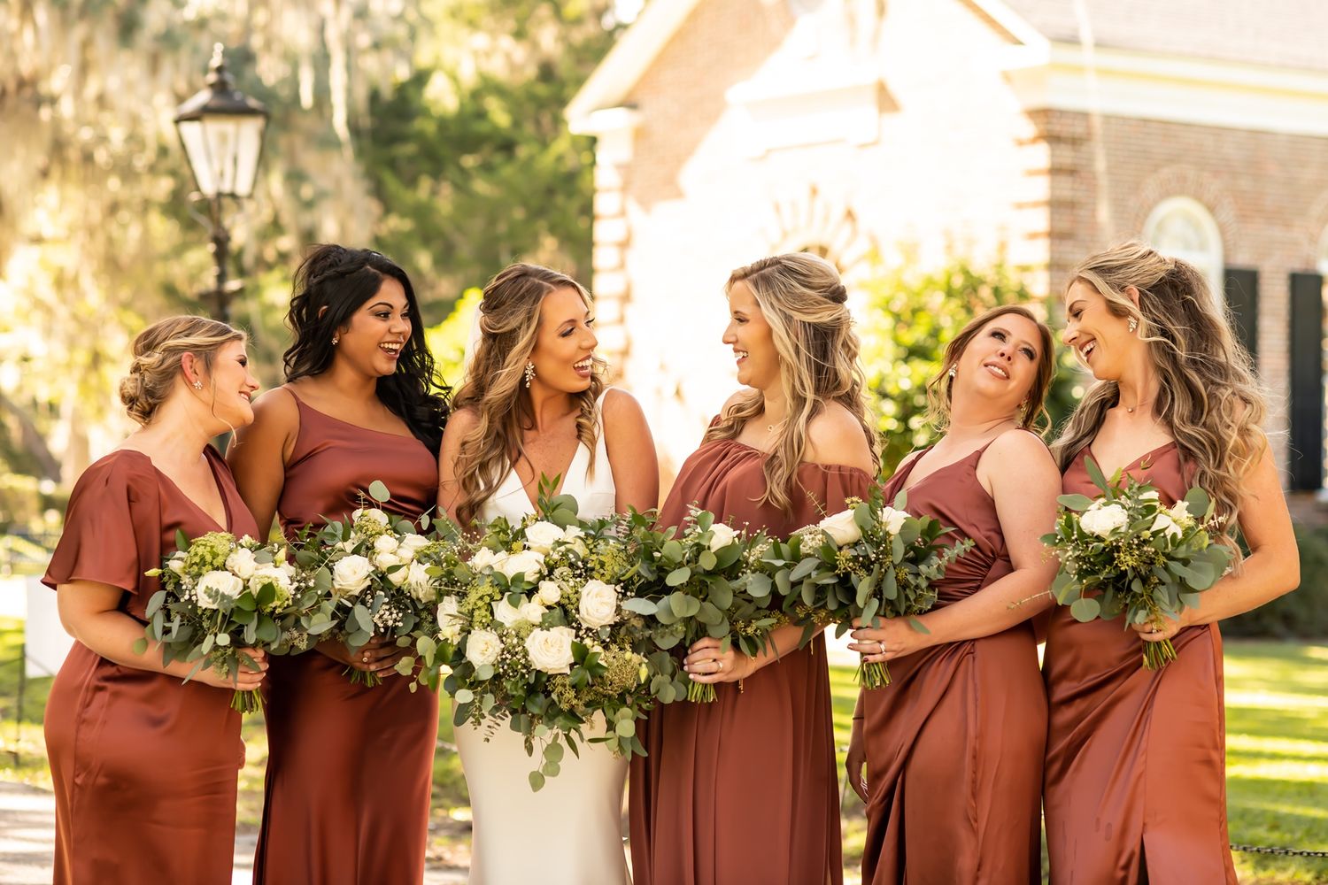 Bridesmaids in rust-colored dresses laugh together while holding white and green floral bouquets outdoors.