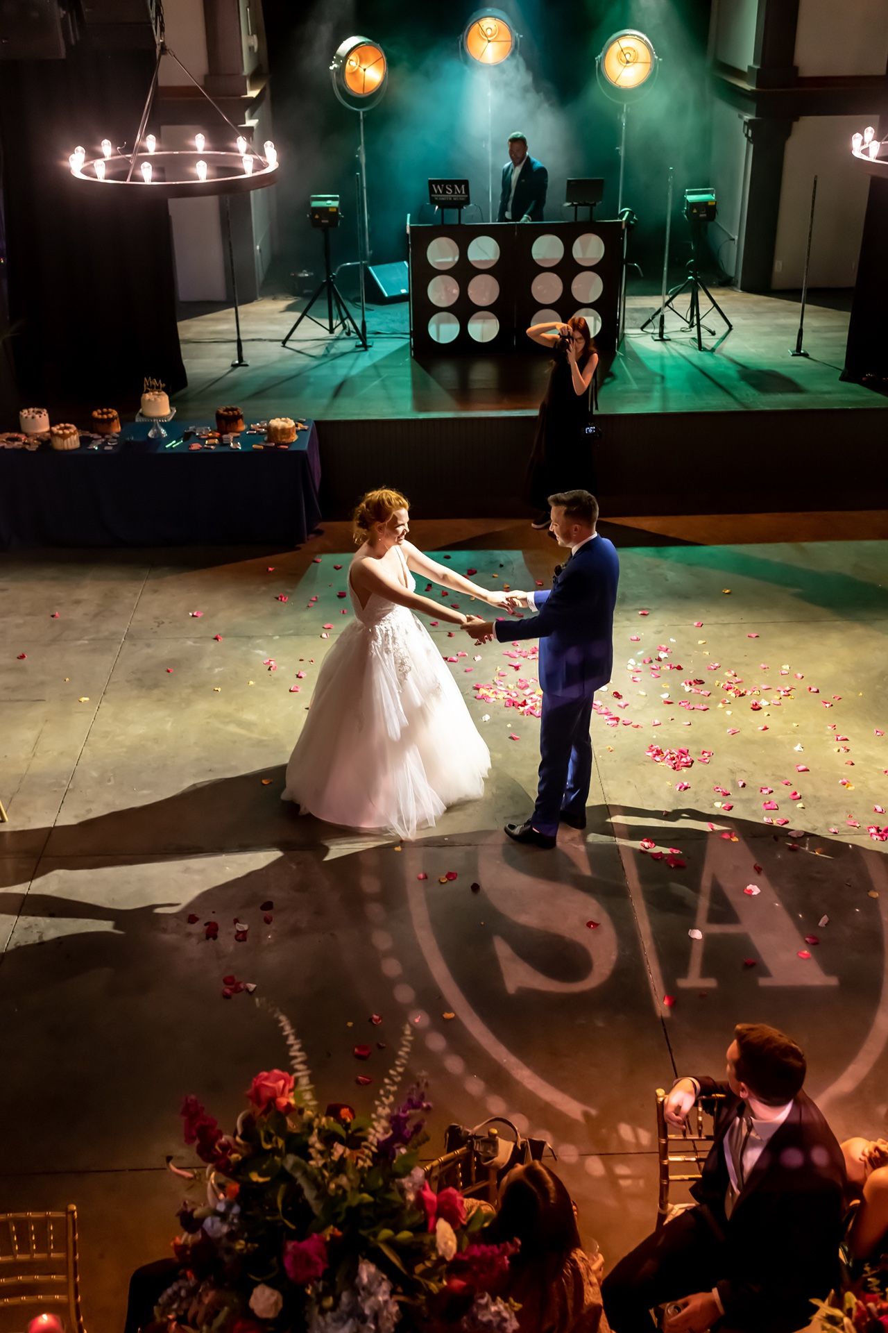 Couple performs romantic first dance at wedding reception under dim lighting with scattered rose petals on the floor.