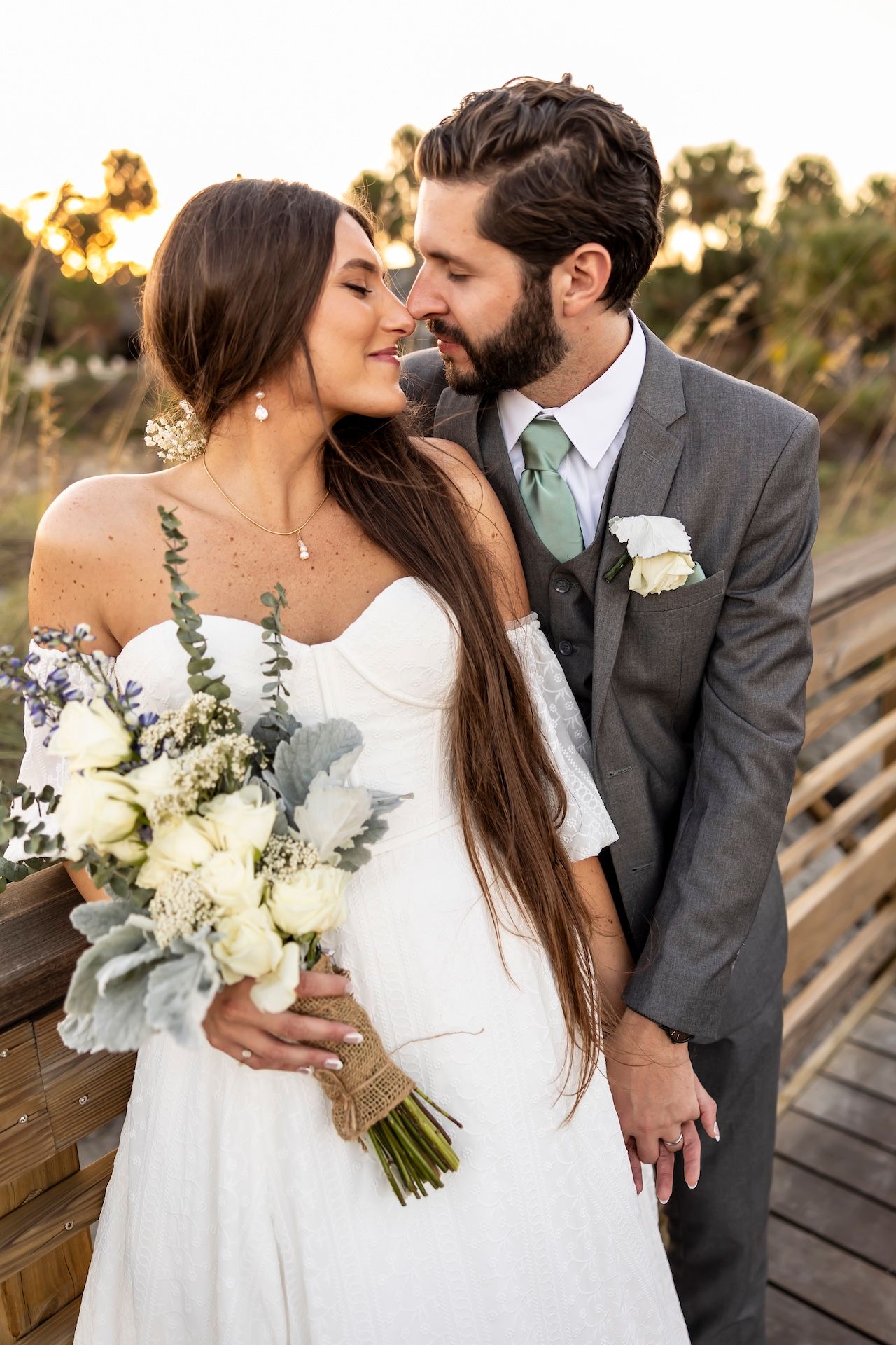 Couple in wedding attire share an intimate moment at sunset with elegant white floral bouquet on wooden boardwalk.