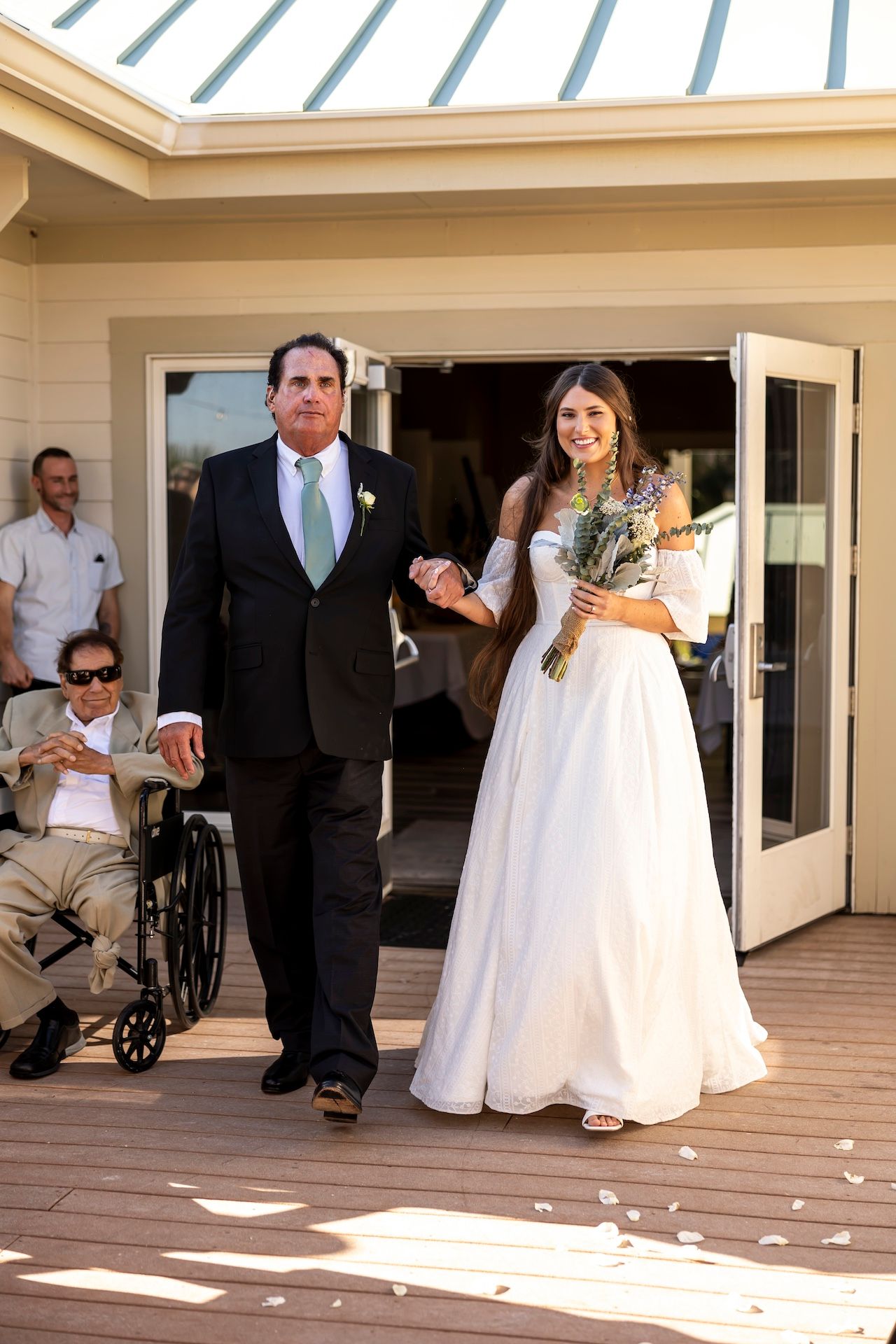 Formal couple exits building onto sunny deck during wedding celebration with guests watching nearby.