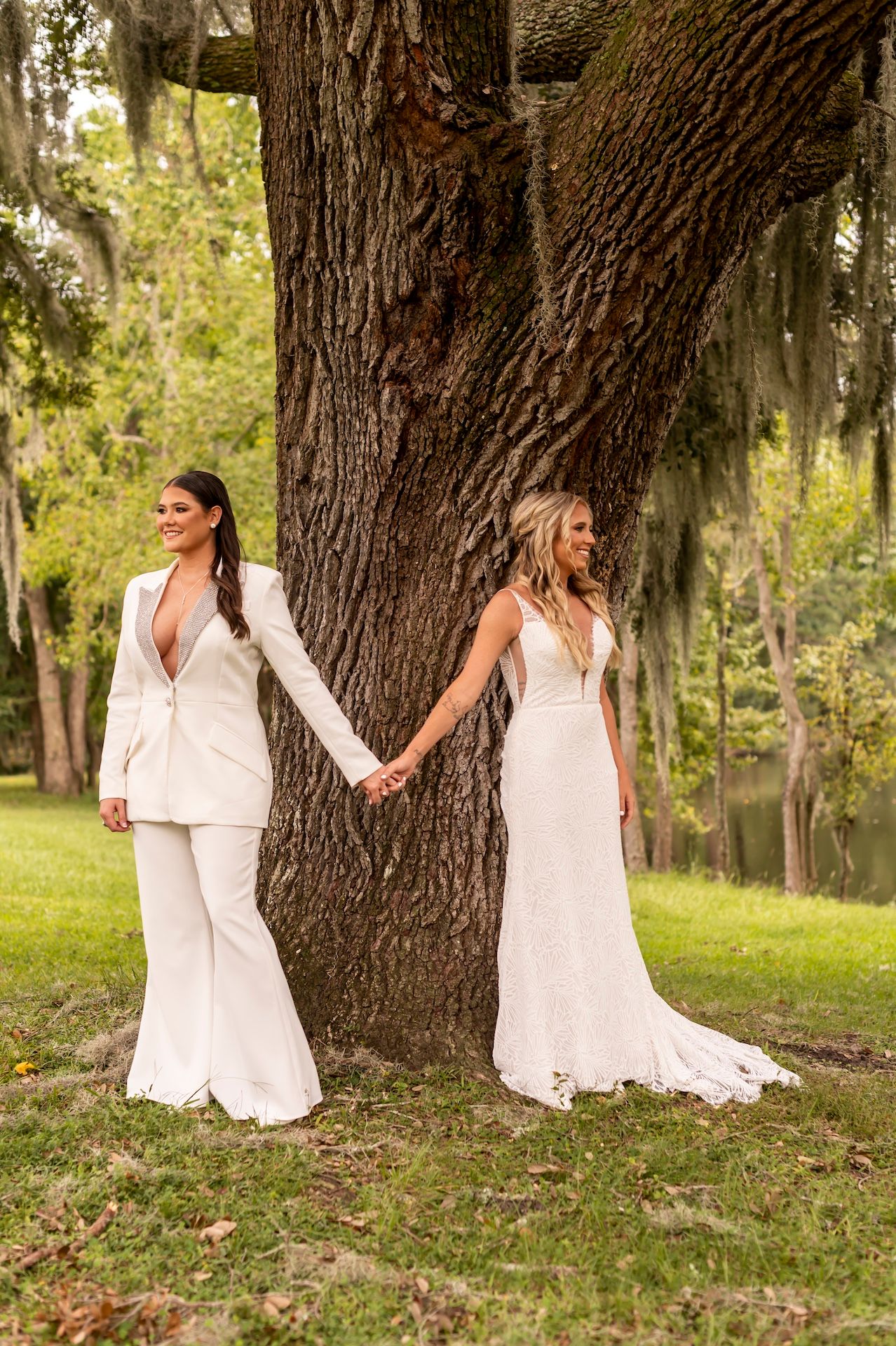 Two people in white outfits holding hands beside a large tree trunk in an outdoor setting with Spanish moss.