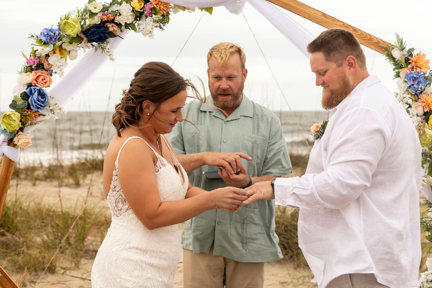 A wedding ceremony takes place under a floral arch on a beach with participants exchanging rings.
