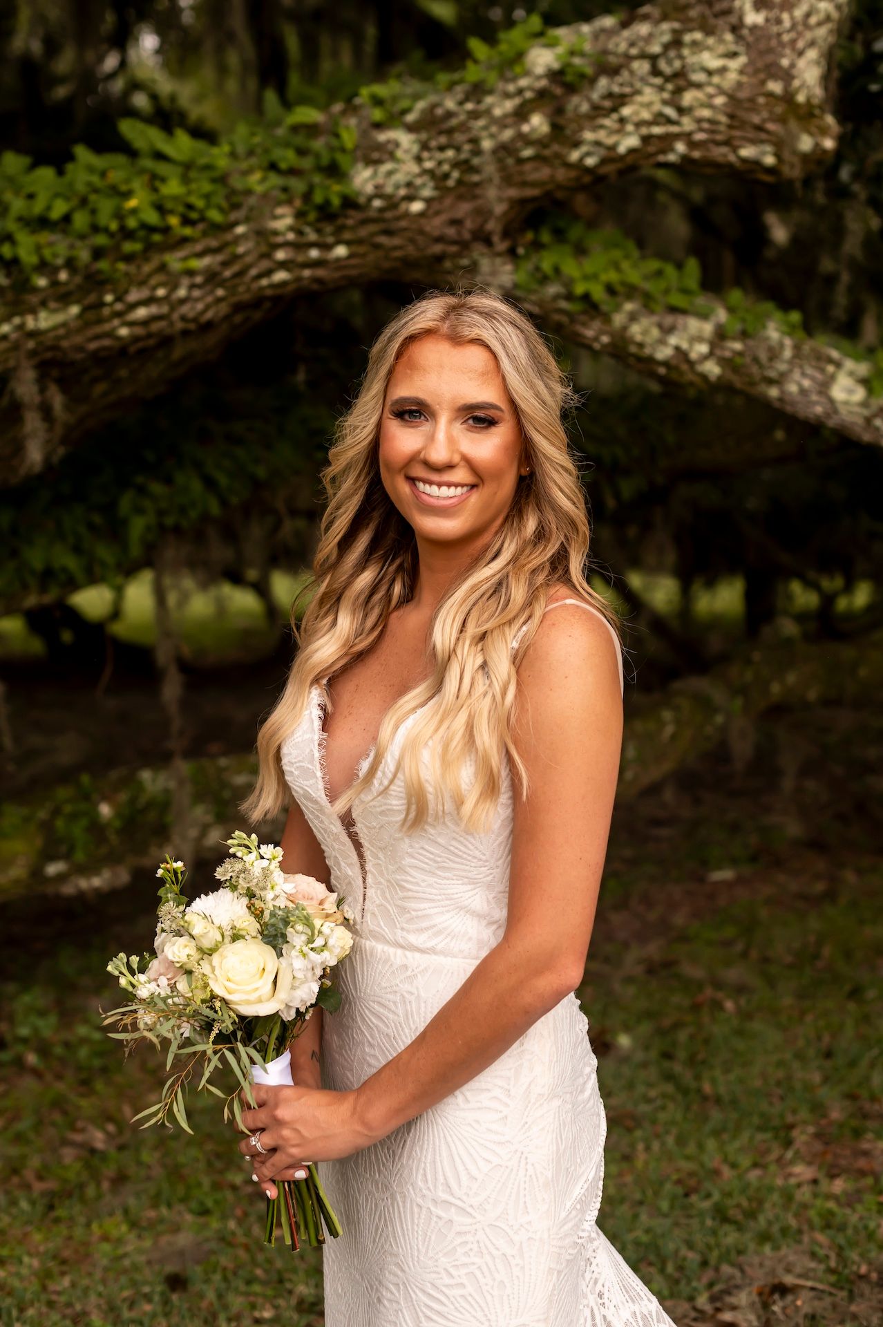 Bride in lace fitted wedding dress holds white bouquet while standing outdoors near natural arch formation.