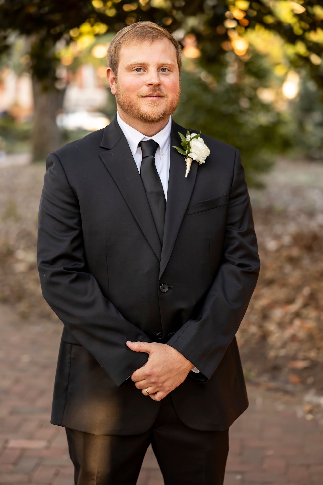 A groom in a classic black suit and tie wears a white boutonniere while standing outdoors on a brick walkway.