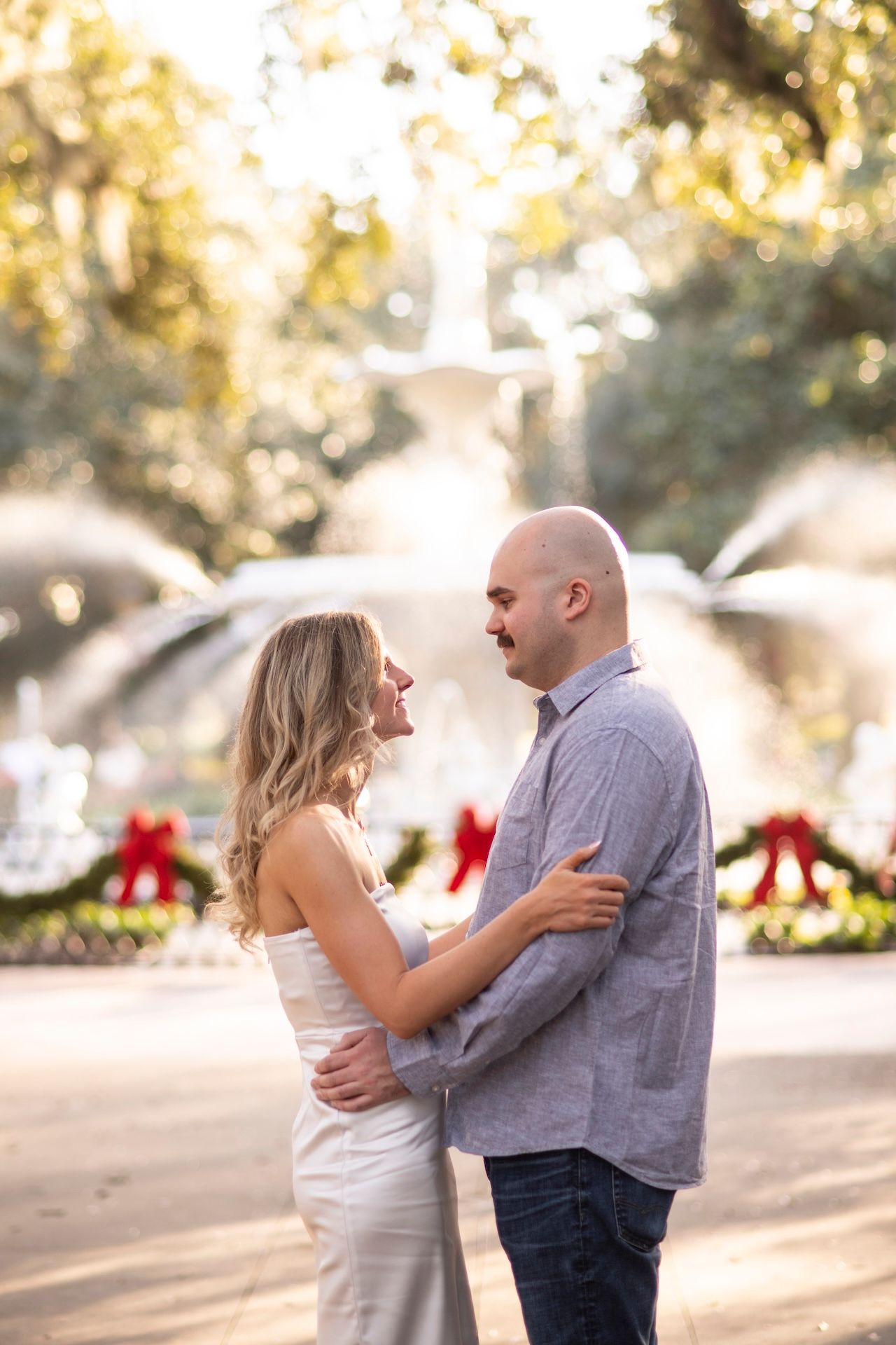A couple shares a romantic moment in front of a fountain surrounded by gardens and holiday decorations at sunset.