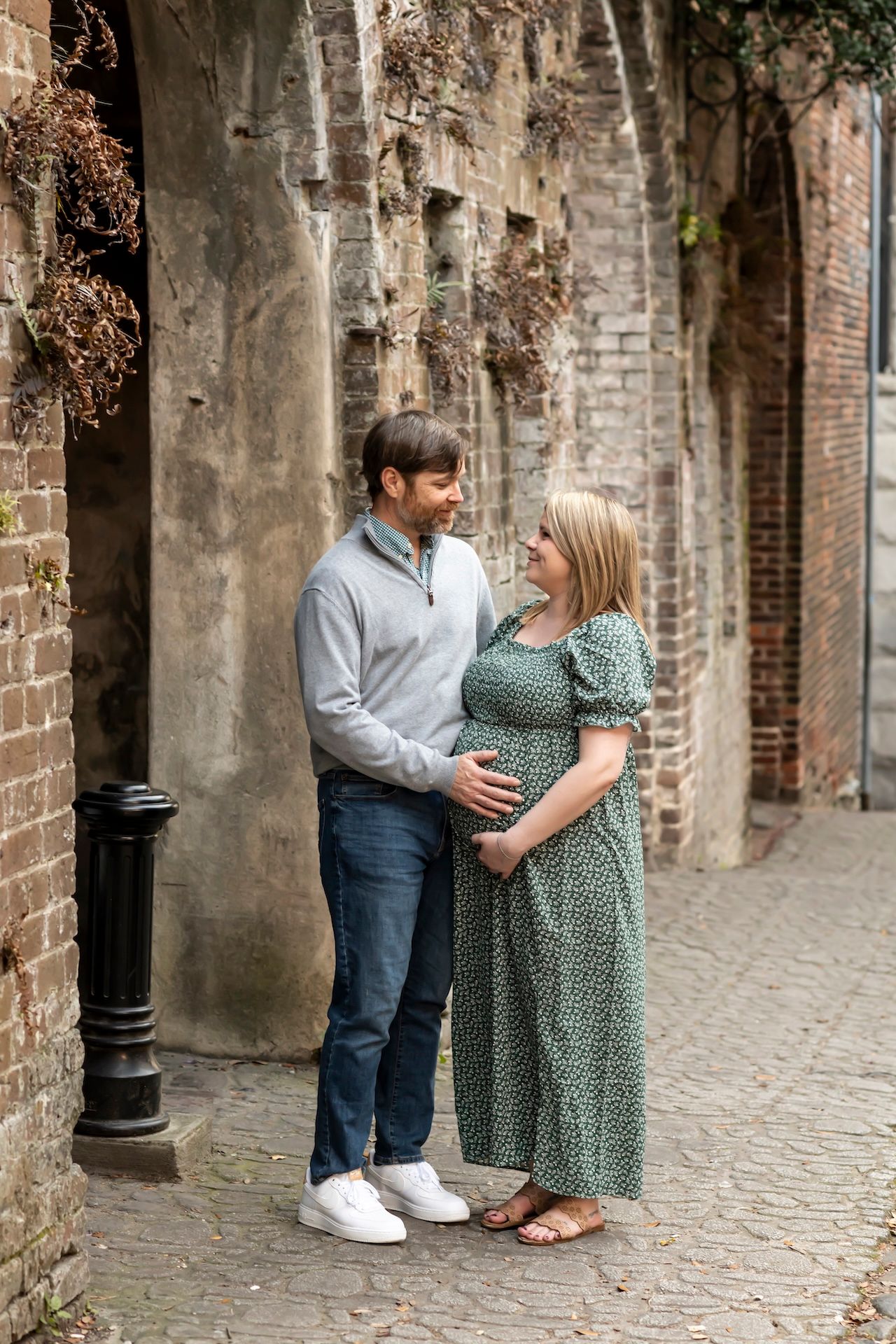 A couple shares a tender moment in a historic brick archway setting, with the expectant woman wearing a green floral dress.