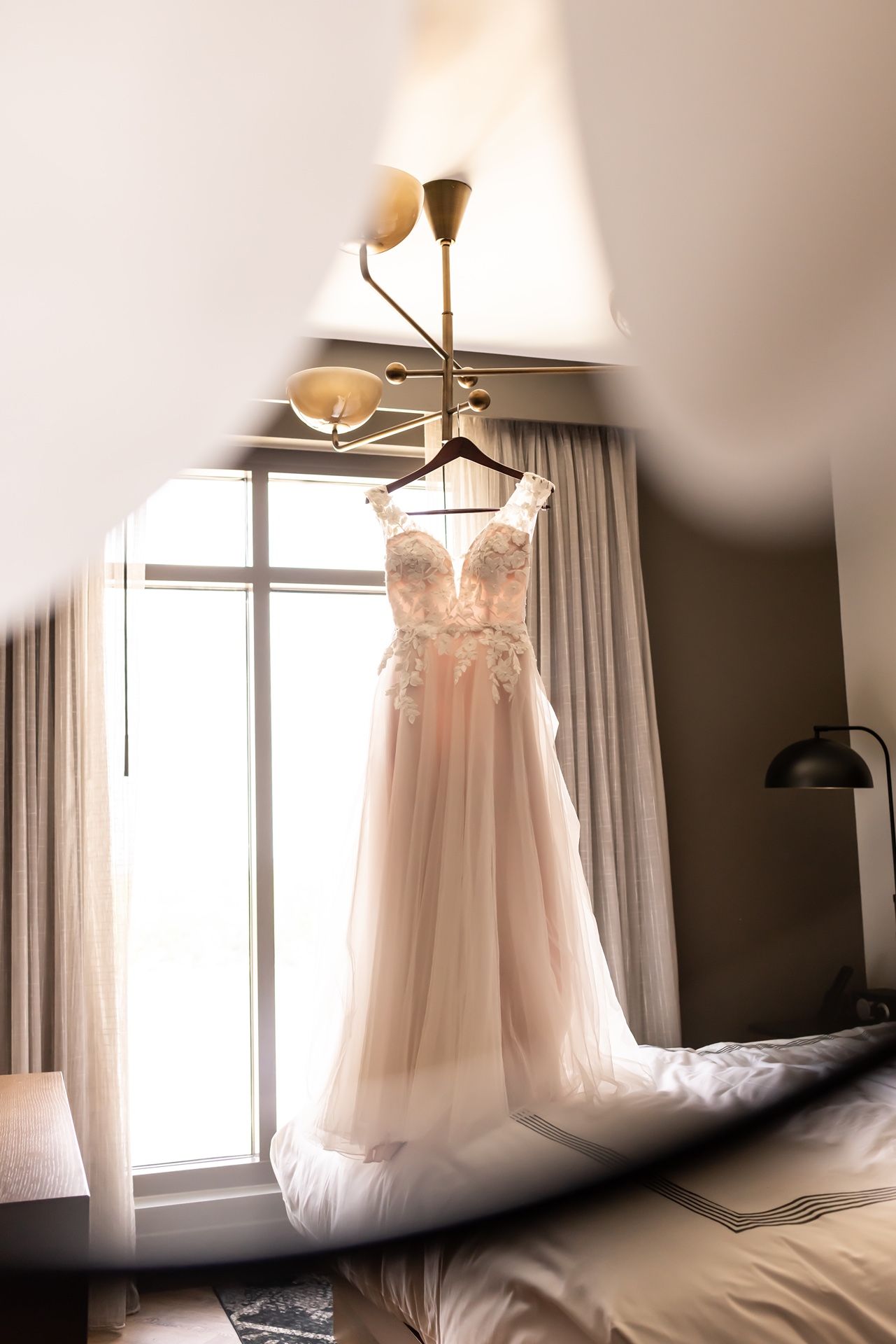 A delicate blush pink wedding dress with lace details hangs elegantly by a sunlit window in a hotel room.