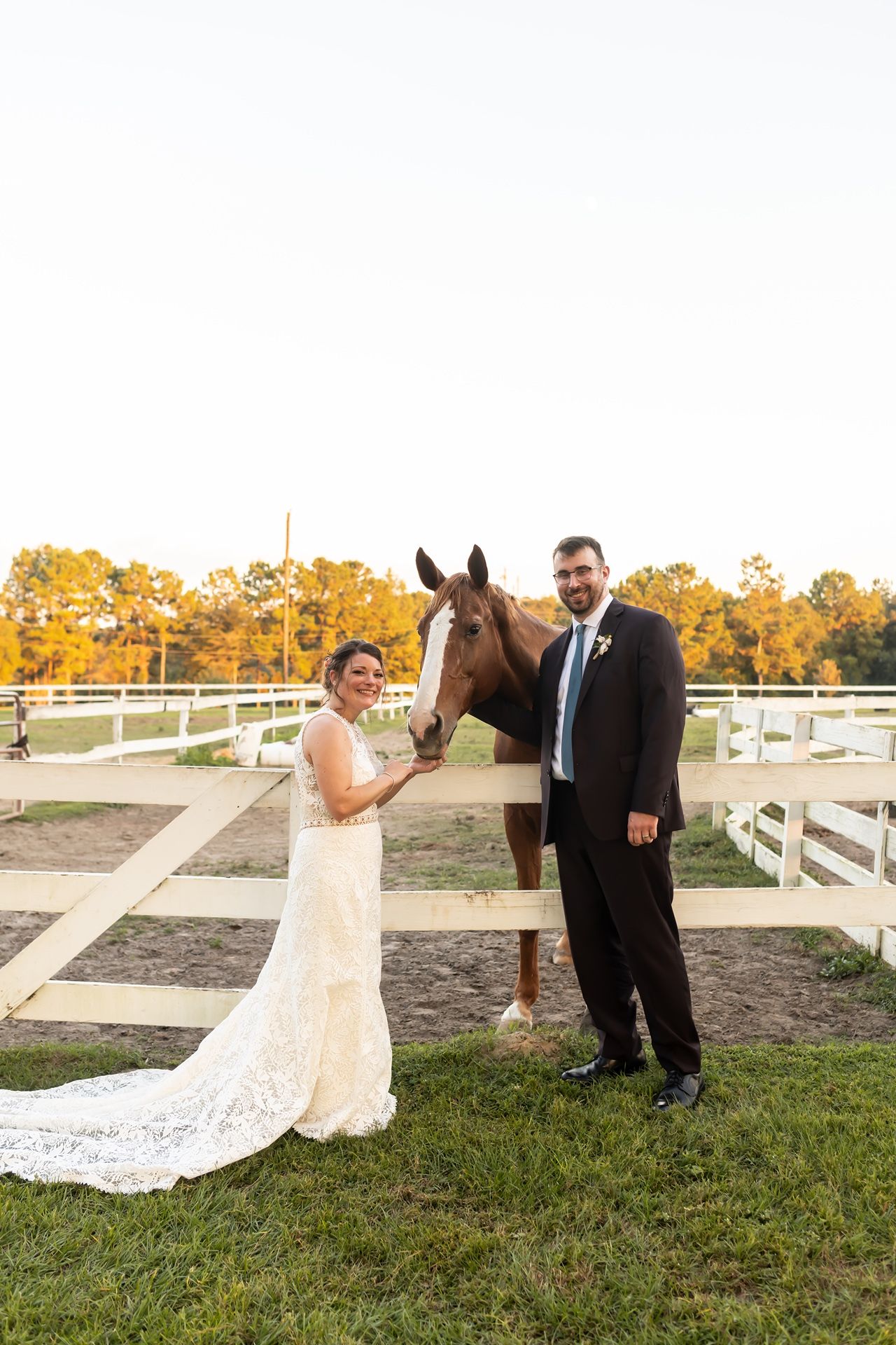 A couple in wedding attire poses with their brown horse near white fencing with autumn trees in the background.