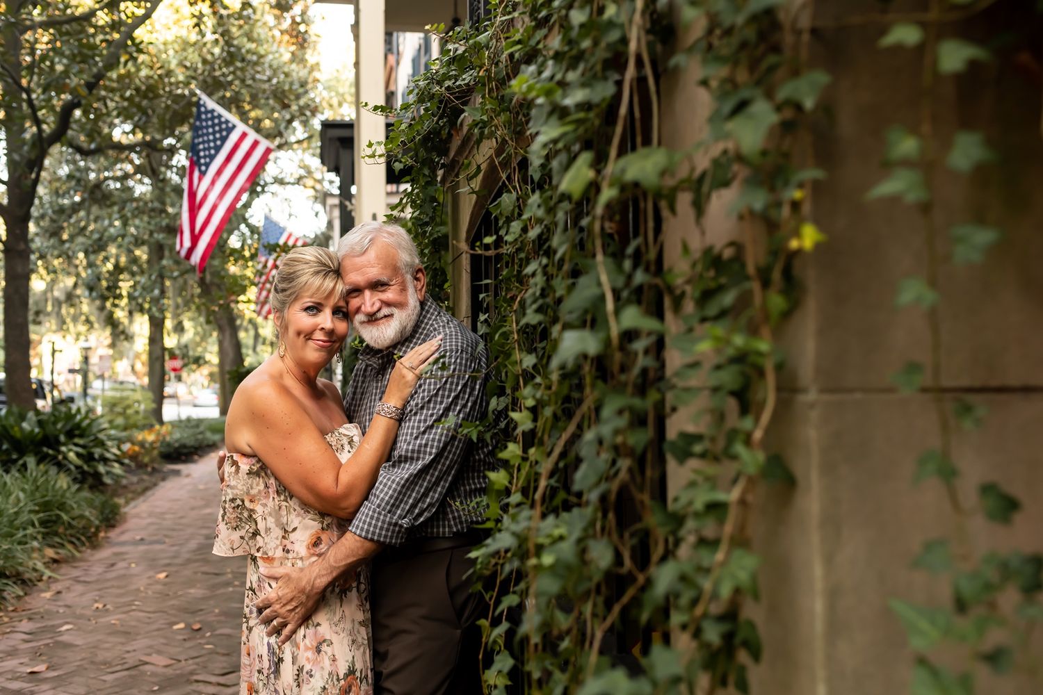 A couple embraces on an ivy-covered porch pathway next to an American flag in a romantic outdoor setting.