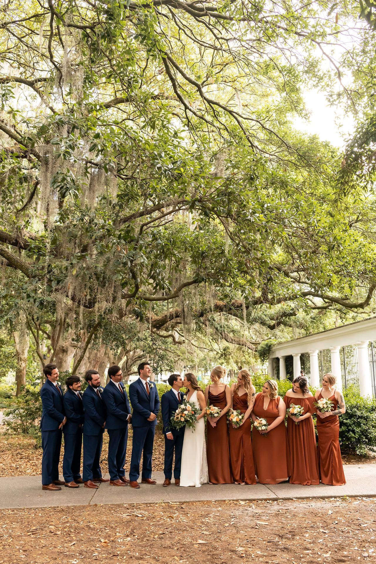 A wedding party stands under majestic Spanish moss-draped oak trees near a white columned building.