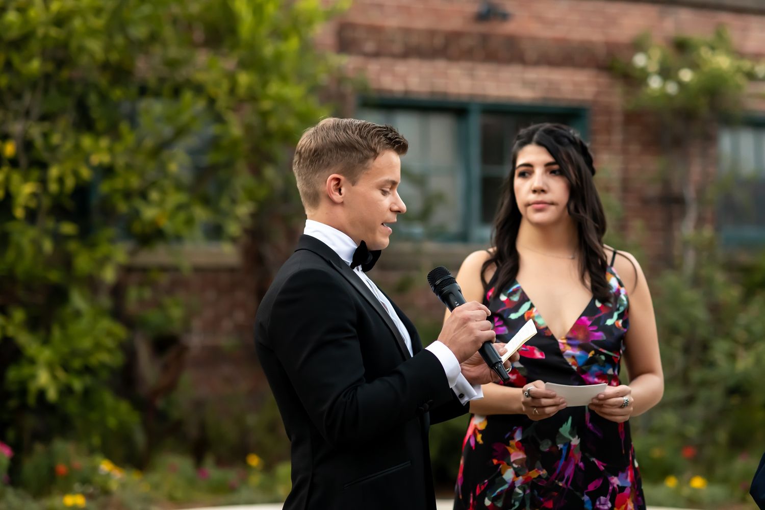 A couple in formal attire stands together in an outdoor garden setting near a brick building.