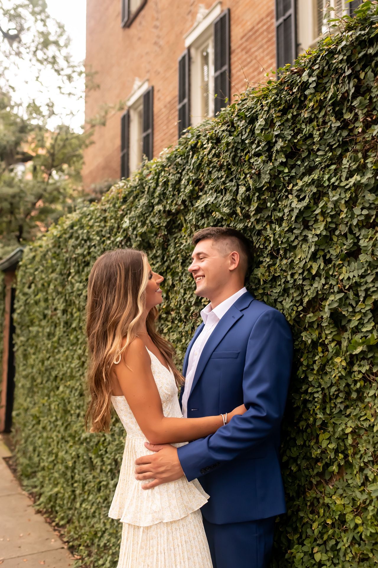 A couple embraces against a brick wall with ivy in an urban setting during a warm afternoon.