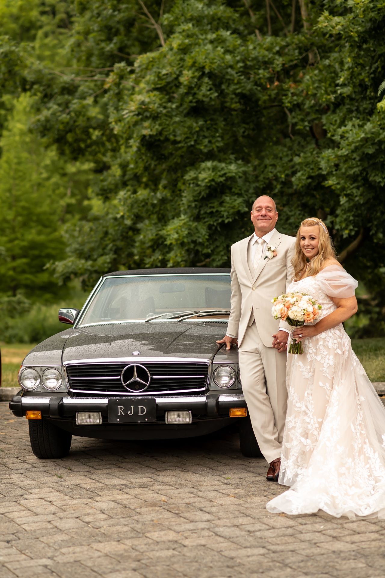 Newlywed couple poses with classic black Mercedes-Benz on cobblestone driveway during outdoor wedding photography session.