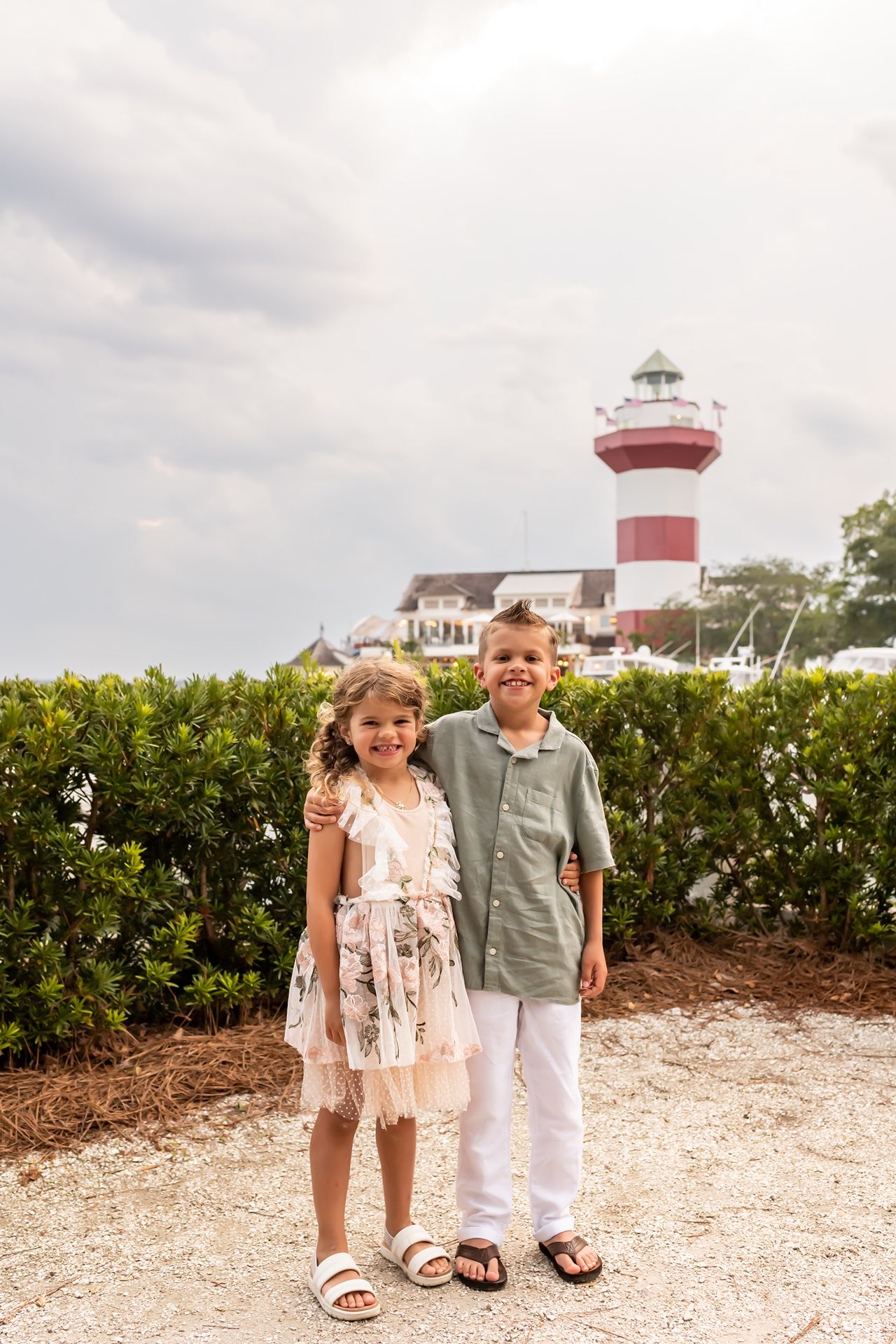 Two children walk and smile along a coastal path with a lighthouse in the background.