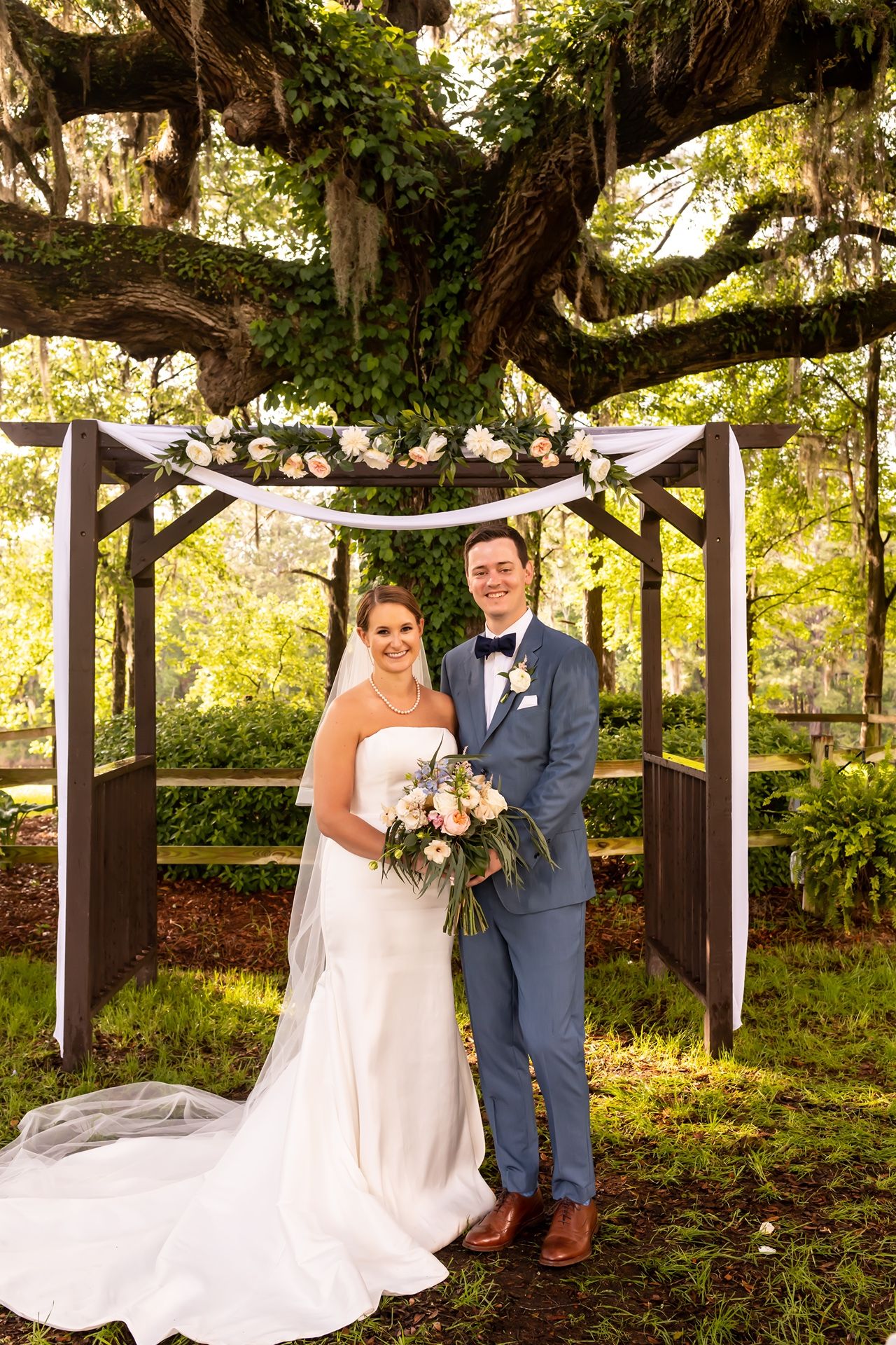 A couple stands under a wooden wedding arch decorated with white flowers and draping fabric in a lush garden setting.