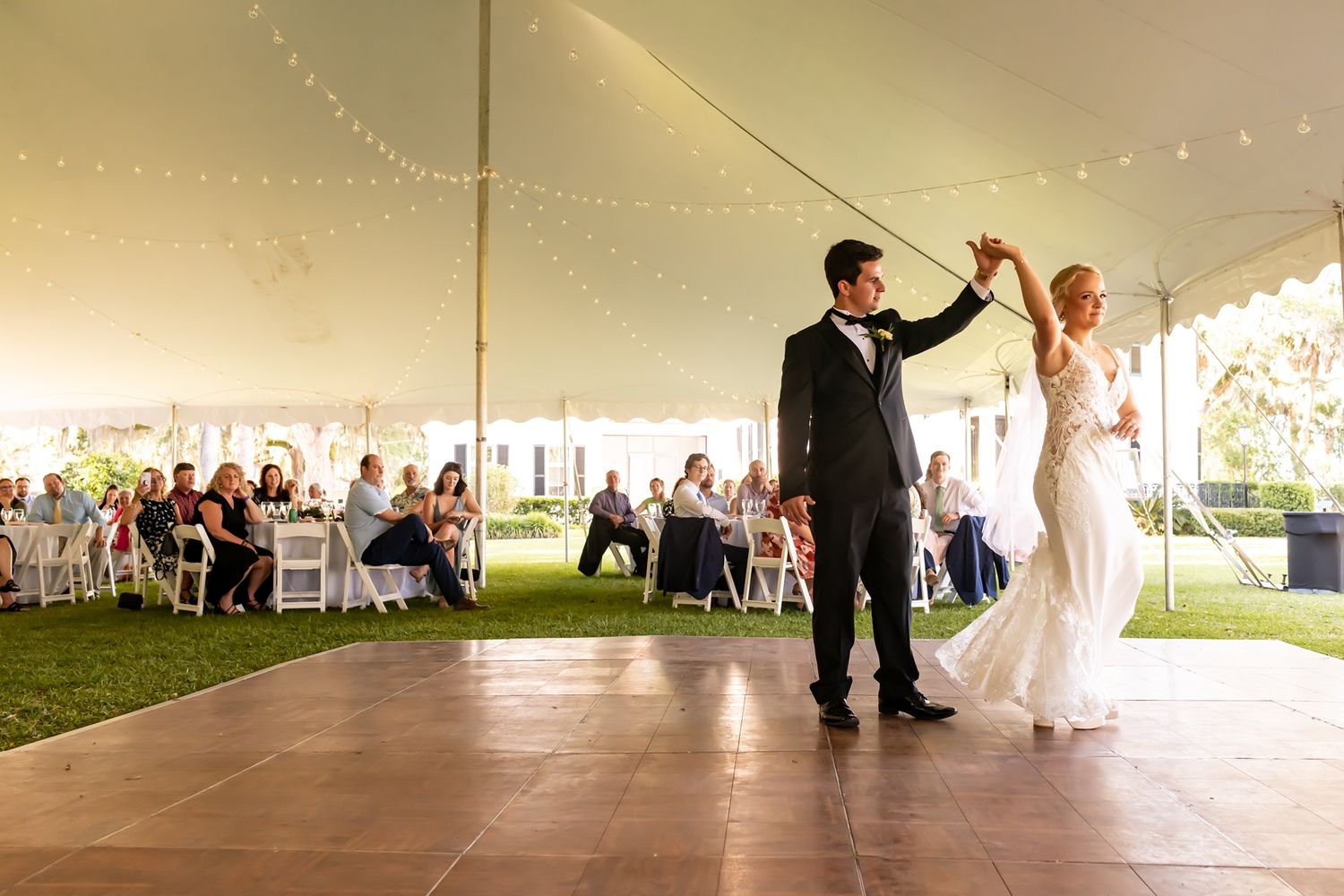 A newlywed couple performing their first dance under a large white tent decorated with string lights.