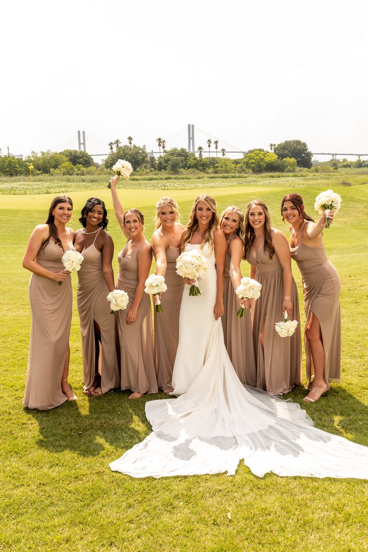 A wedding party in taupe bridesmaid dresses poses for photos on a green lawn with skyline in background.