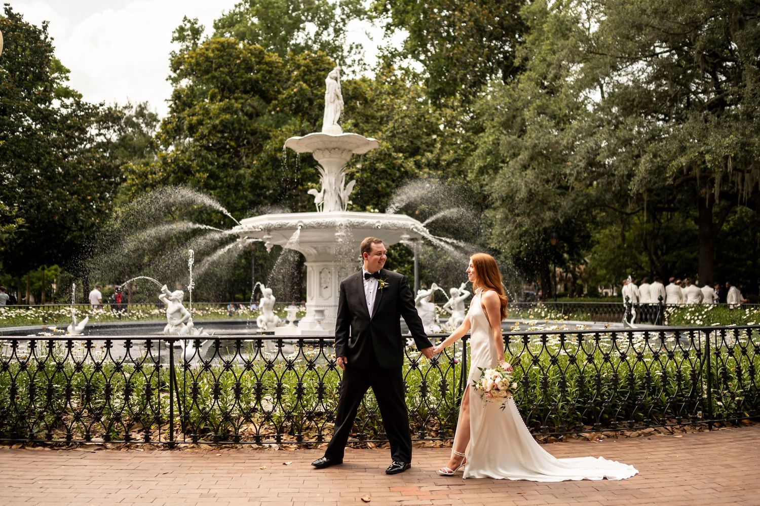 A couple in formal wedding attire poses together in front of an ornate fountain in a lush garden setting.
