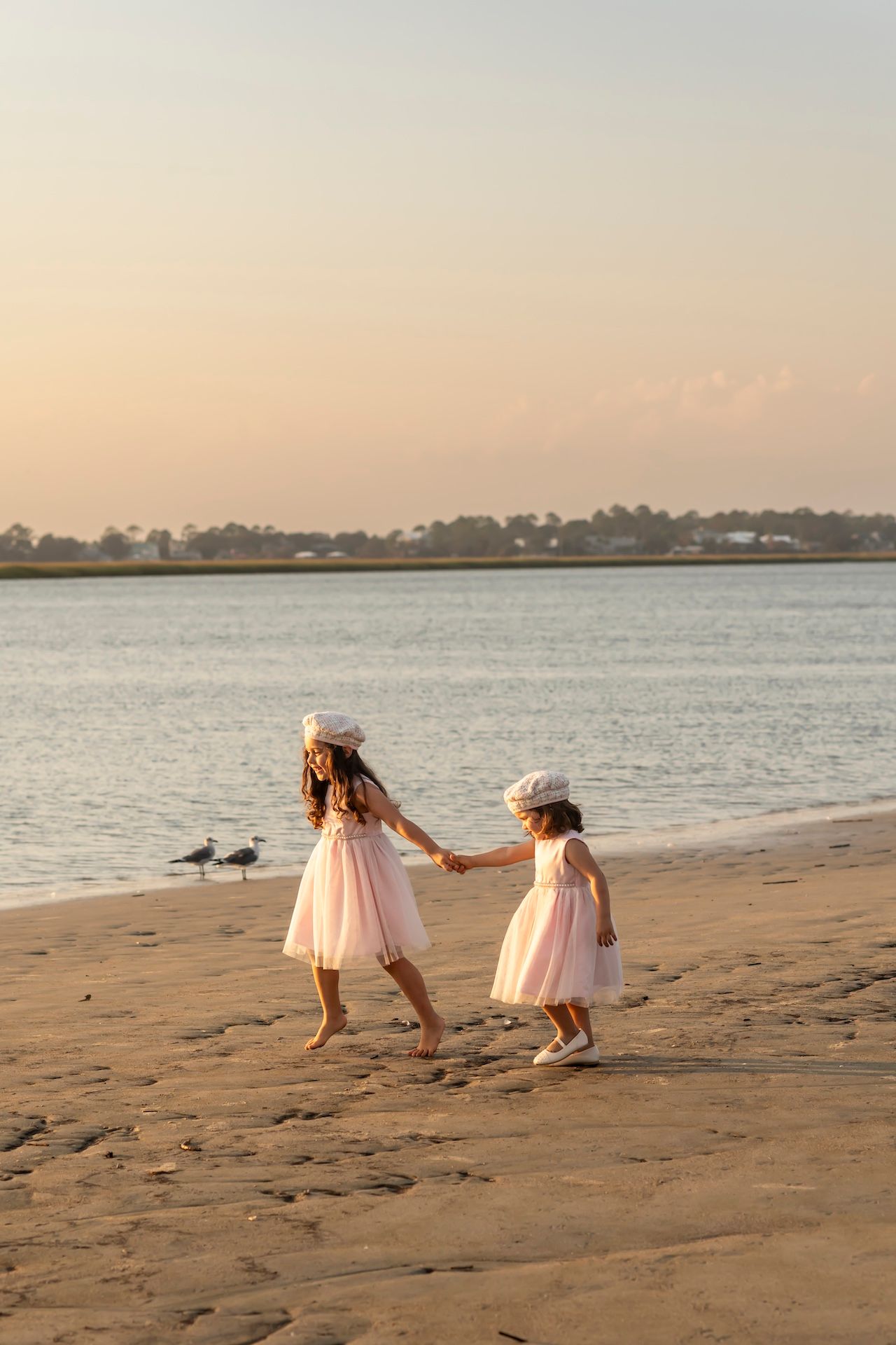 Two young children in peach dresses dance together on a sandy beach at sunset with calm waters and palm trees in background.