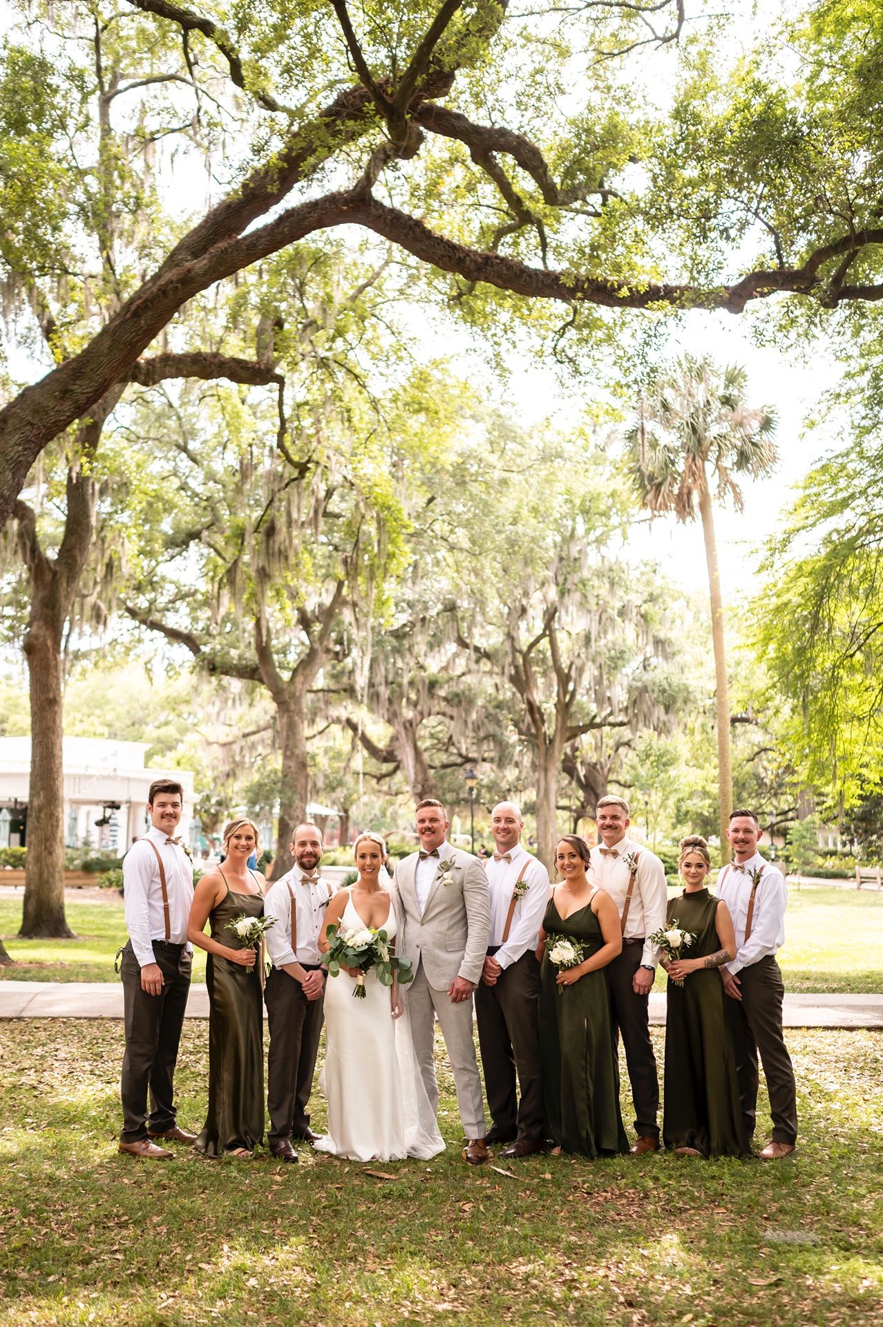 Wedding party in formal attire stands beneath Spanish moss-draped oak trees in a sun-drenched outdoor setting.