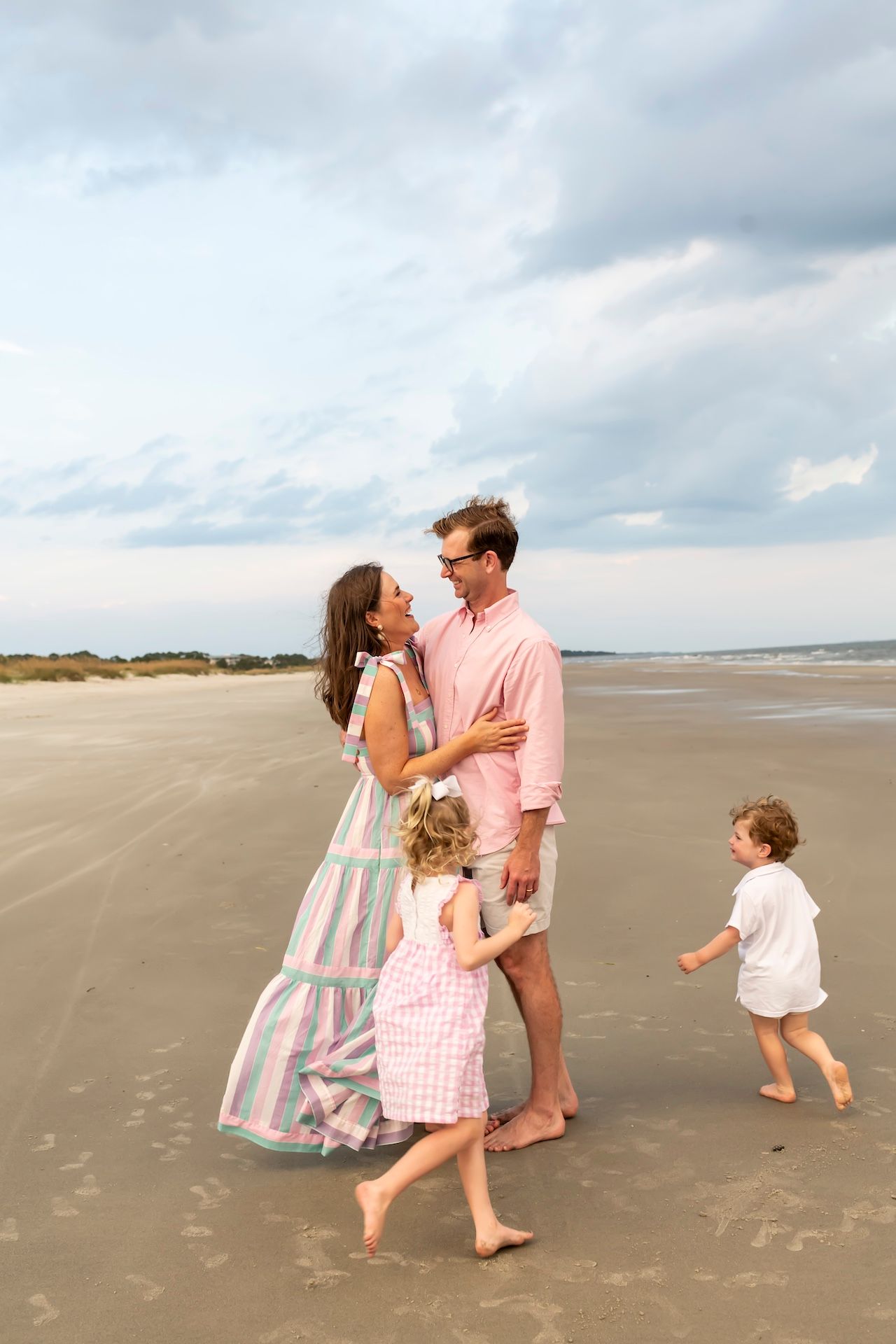 A barefoot family plays and dances together on an expansive beach with cloudy skies overhead.