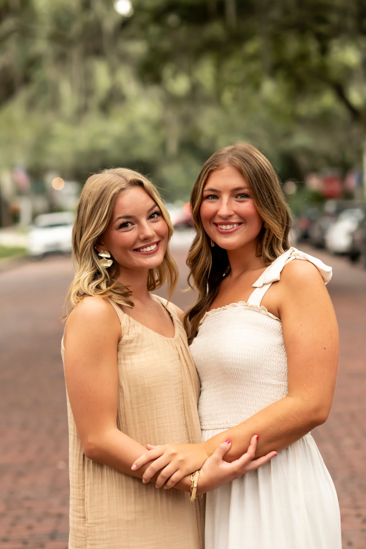 Two women in beige and white dresses pose together on a brick street with trees and cars in the background.
