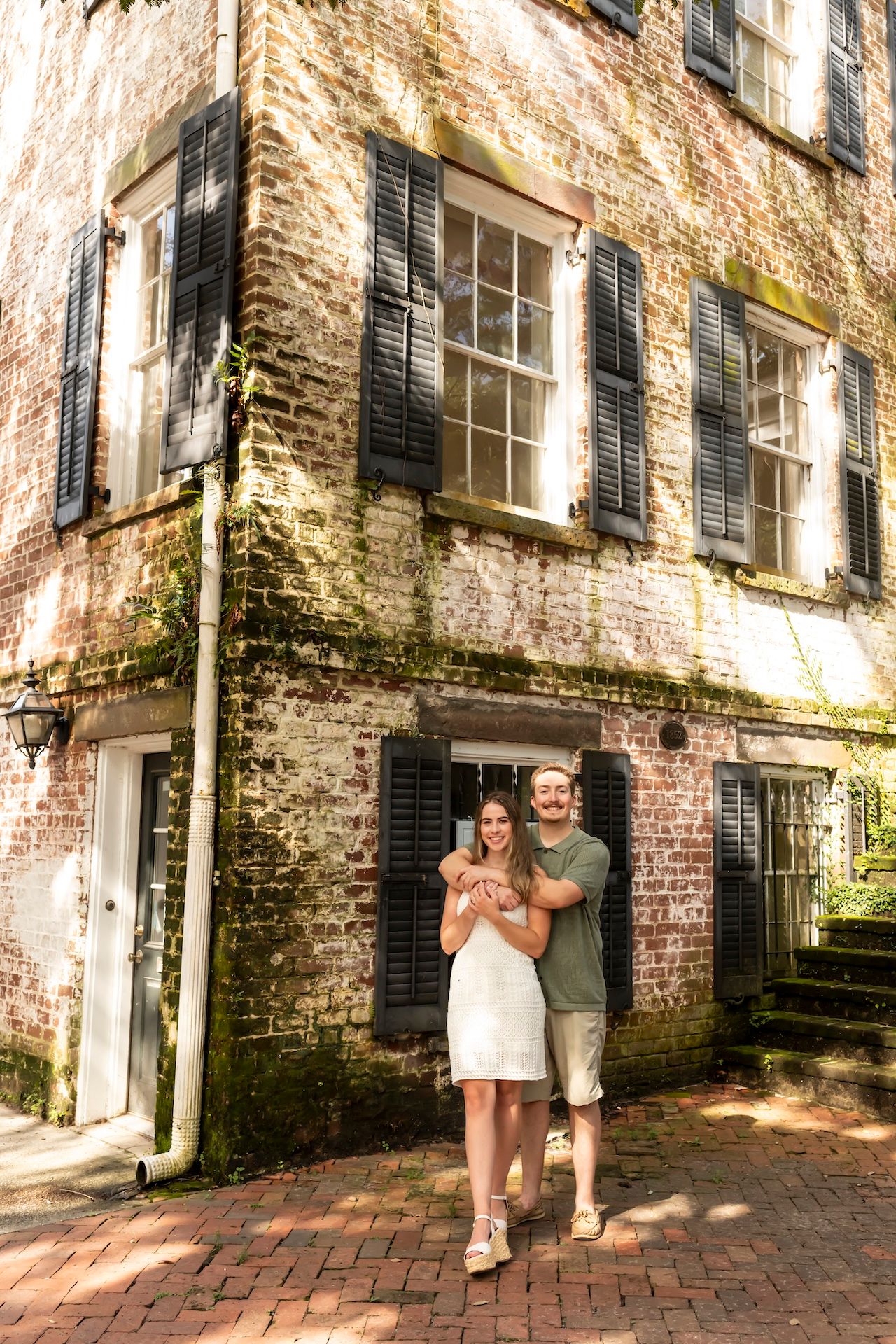 A couple stands outside a historic brick building with dark shutters and moss-covered walls on a brick sidewalk.
