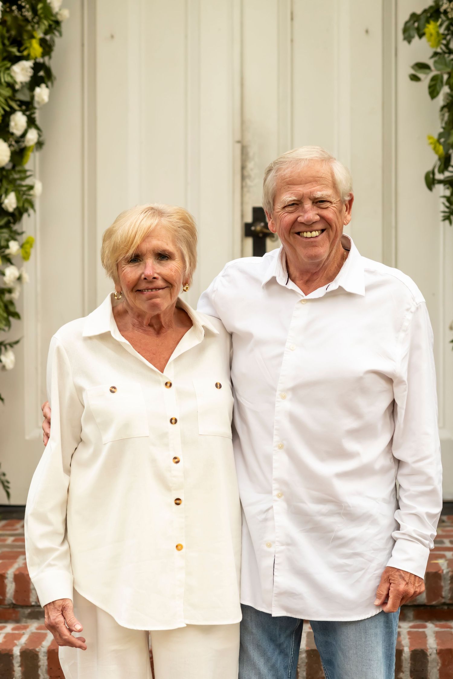 An elderly couple in matching white shirts share a candid laugh while posing against a white wall with green foliage decor.