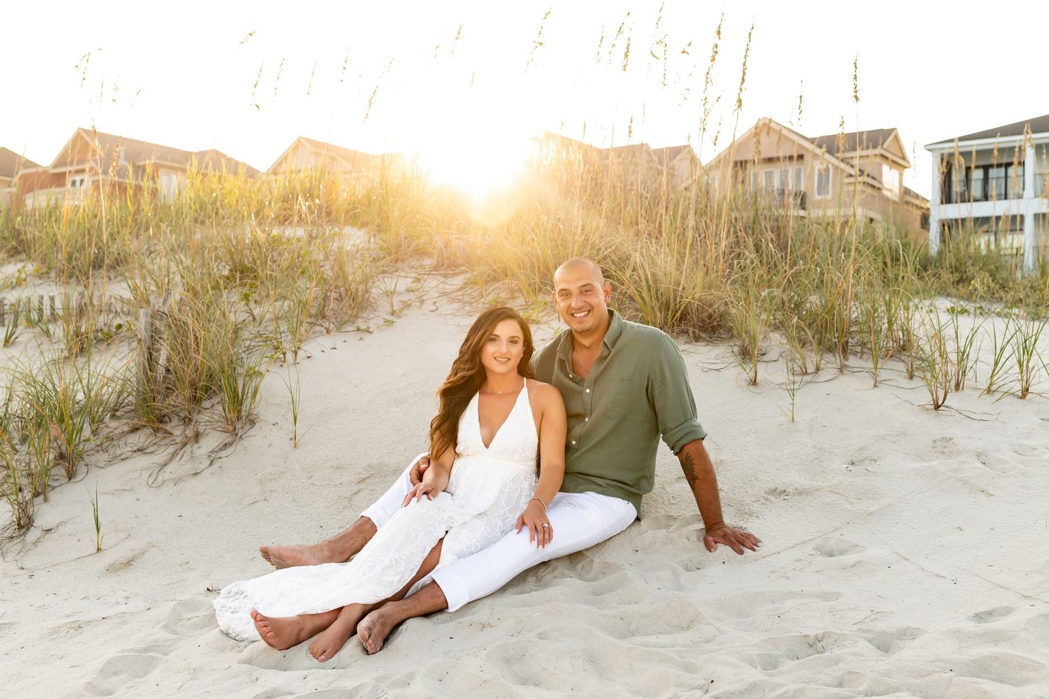 A couple in white clothing poses together on a sandy beach dune at sunset.