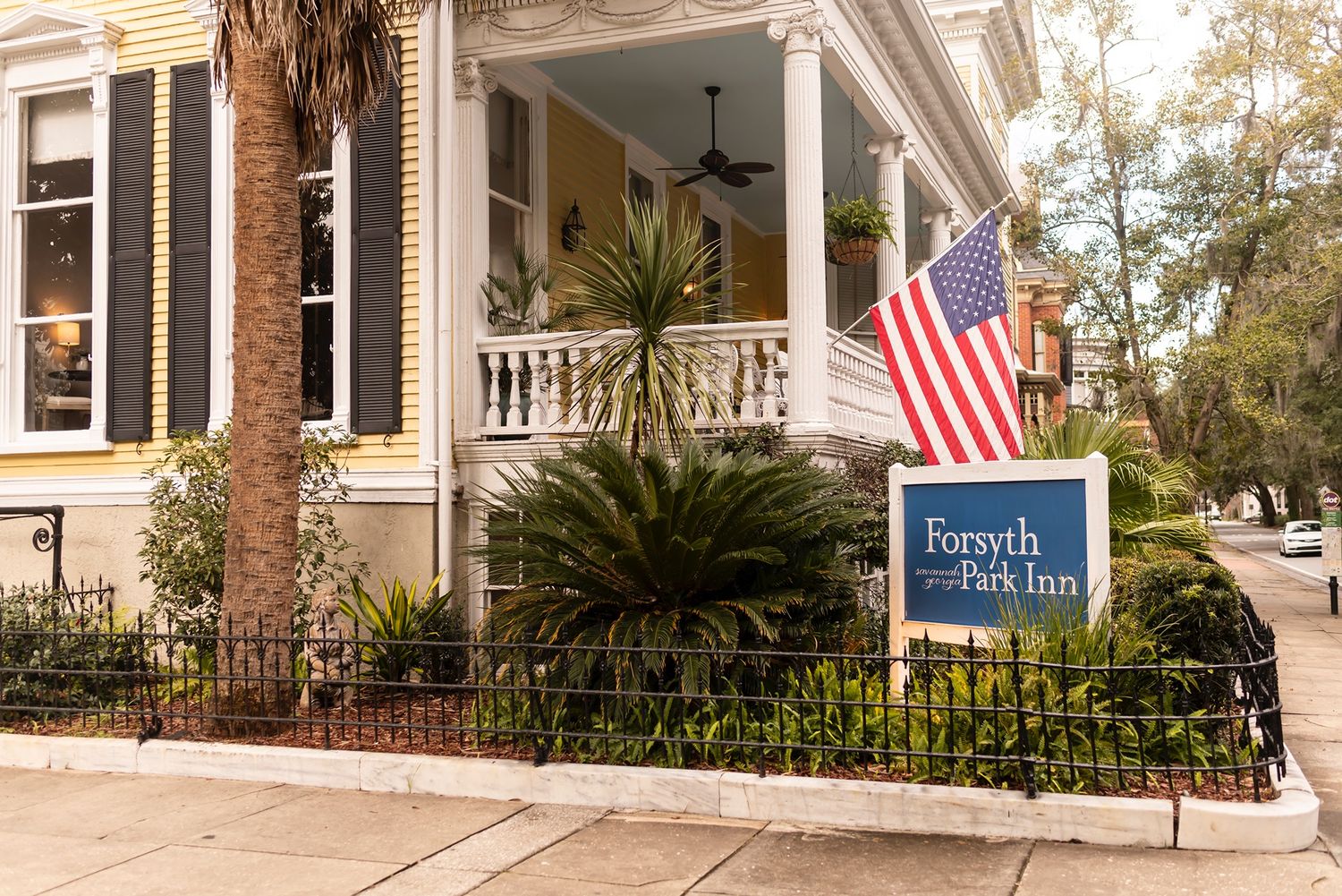 A historic coastal inn with a Southern wraparound porch, American flag, and palm trees in the foreground.