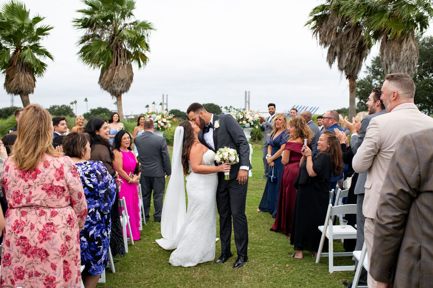 Bride and groom share a kiss at outdoor wedding ceremony with palm trees and guests lining the aisle.