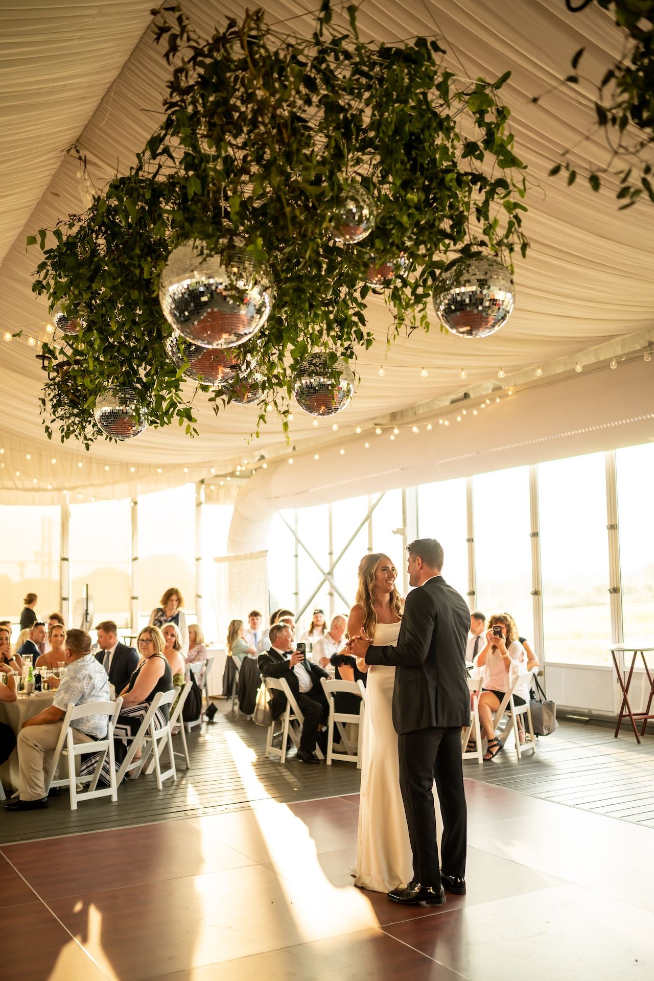Bride and groom share first dance under hanging greenery and disco balls at elegant wedding reception.