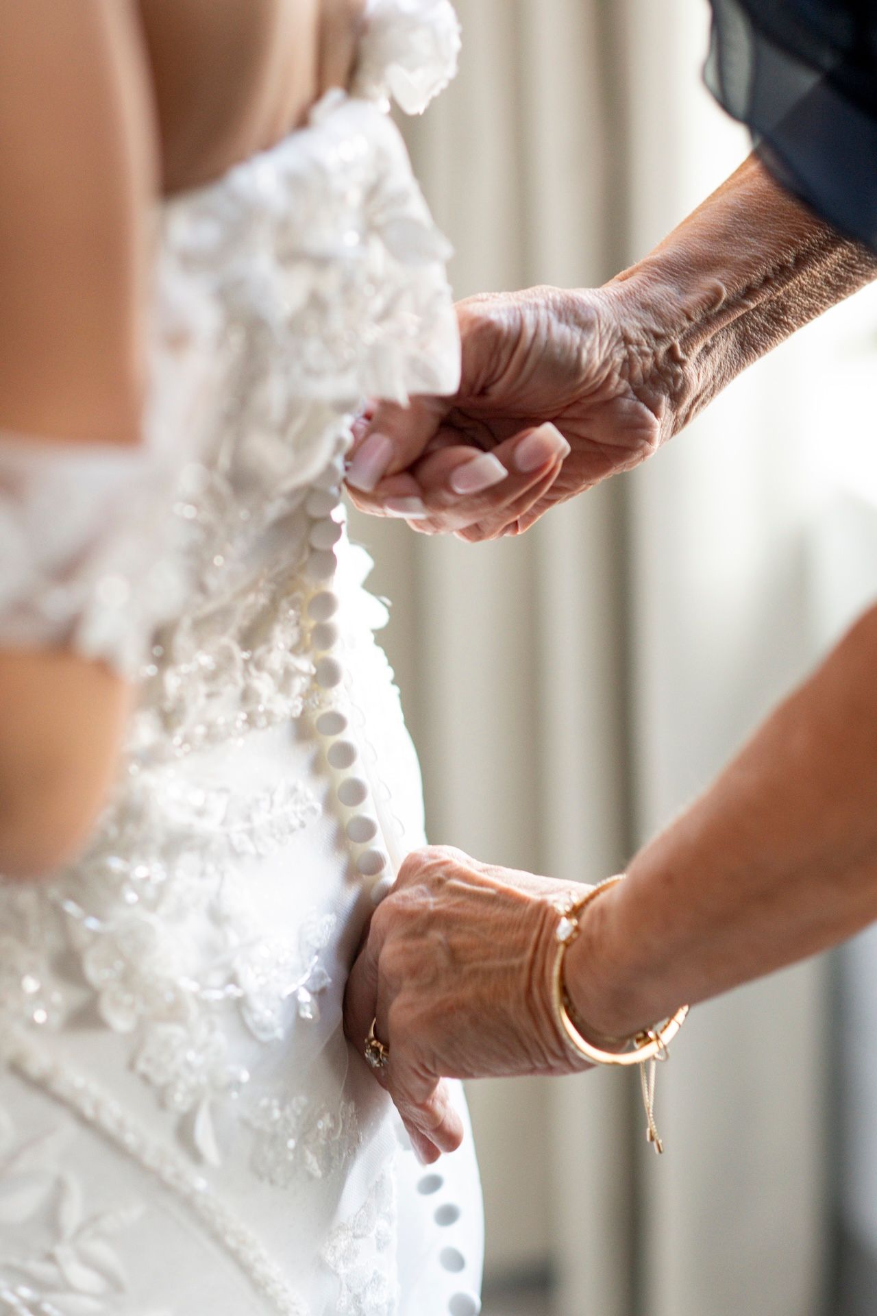 Helping hands fasten delicate buttons on an elegant white lace wedding dress with pearl details.