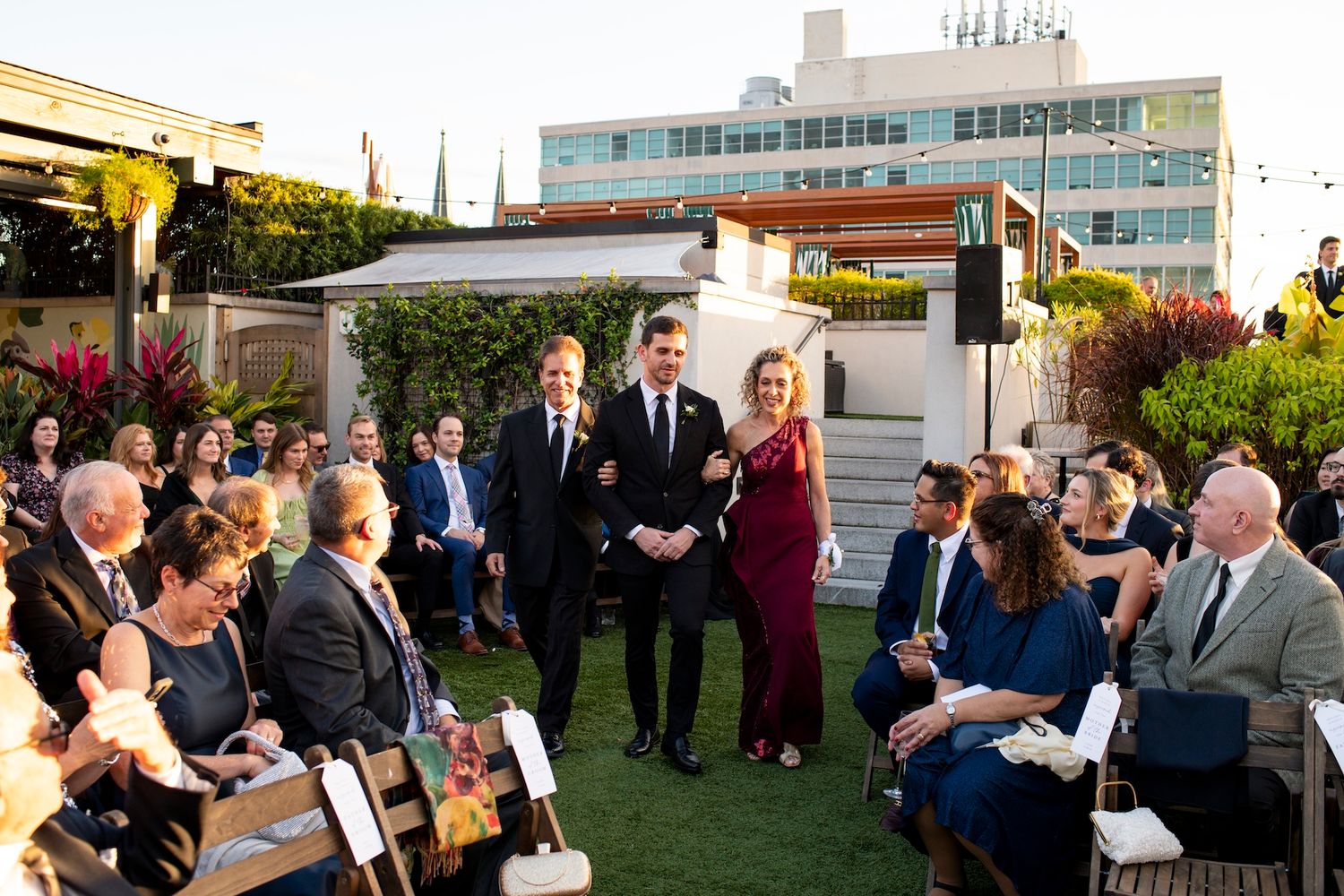 A small outdoor wedding ceremony takes place on a rooftop garden venue at sunset with city buildings in the background.