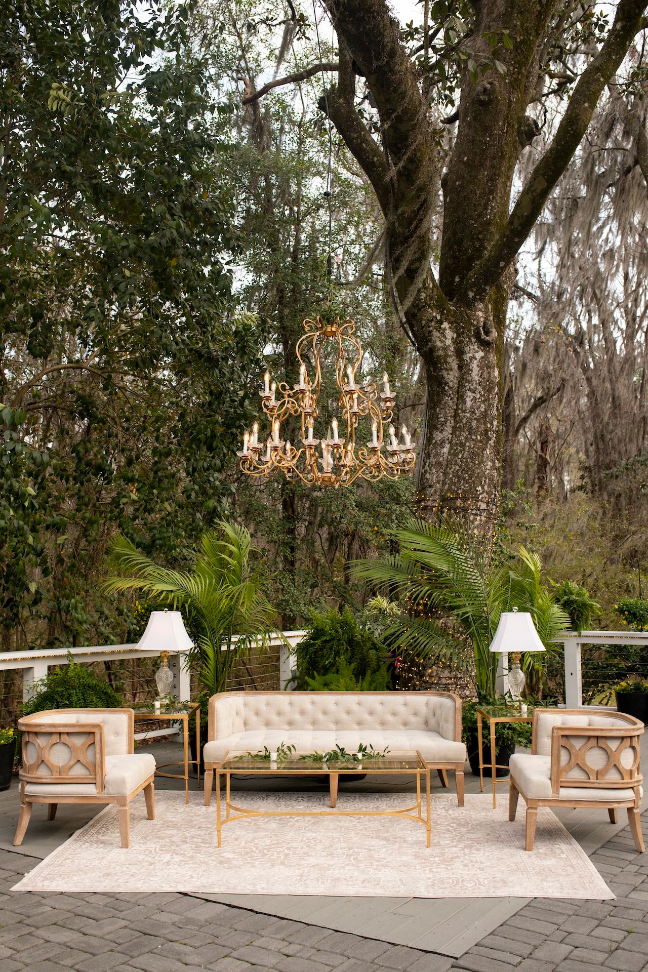 Elegant outdoor lounge with tufted sofa, gold chairs, chandelier, and lush greenery for a wedding reception.