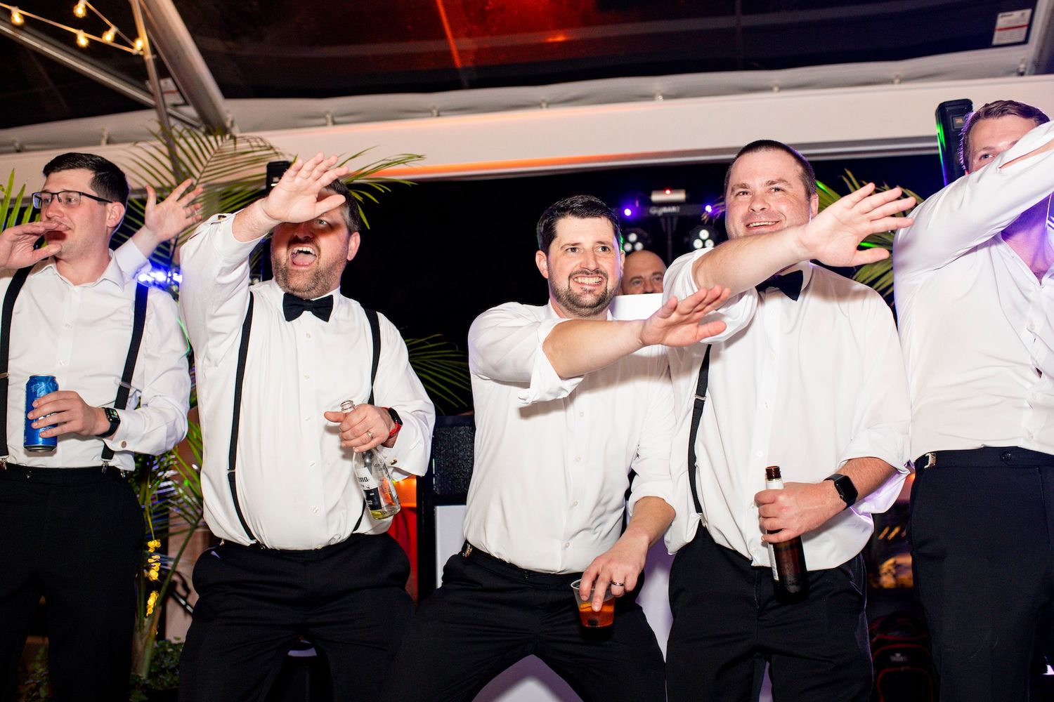 Five groomsmen in white shirts and suspenders celebrating on a dance floor at a wedding reception.