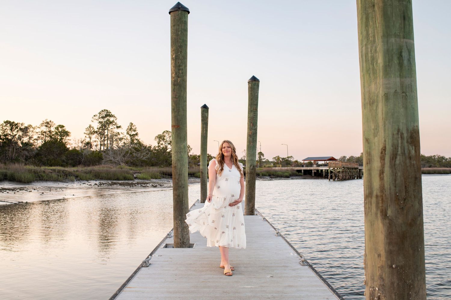 Young woman in white floral dress walking on wooden dock at sunset over calm coastal waters.