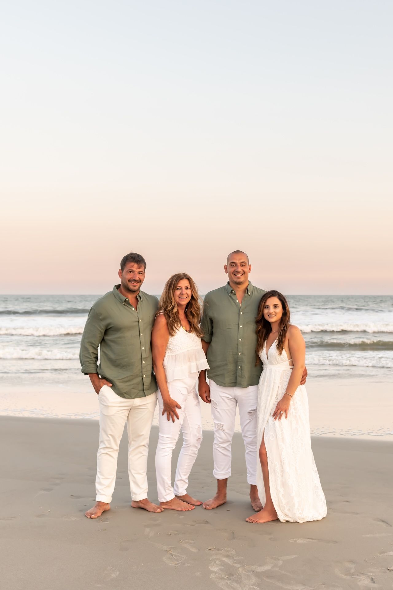 Four people in white and green outfits pose together on a sandy beach at sunset.
