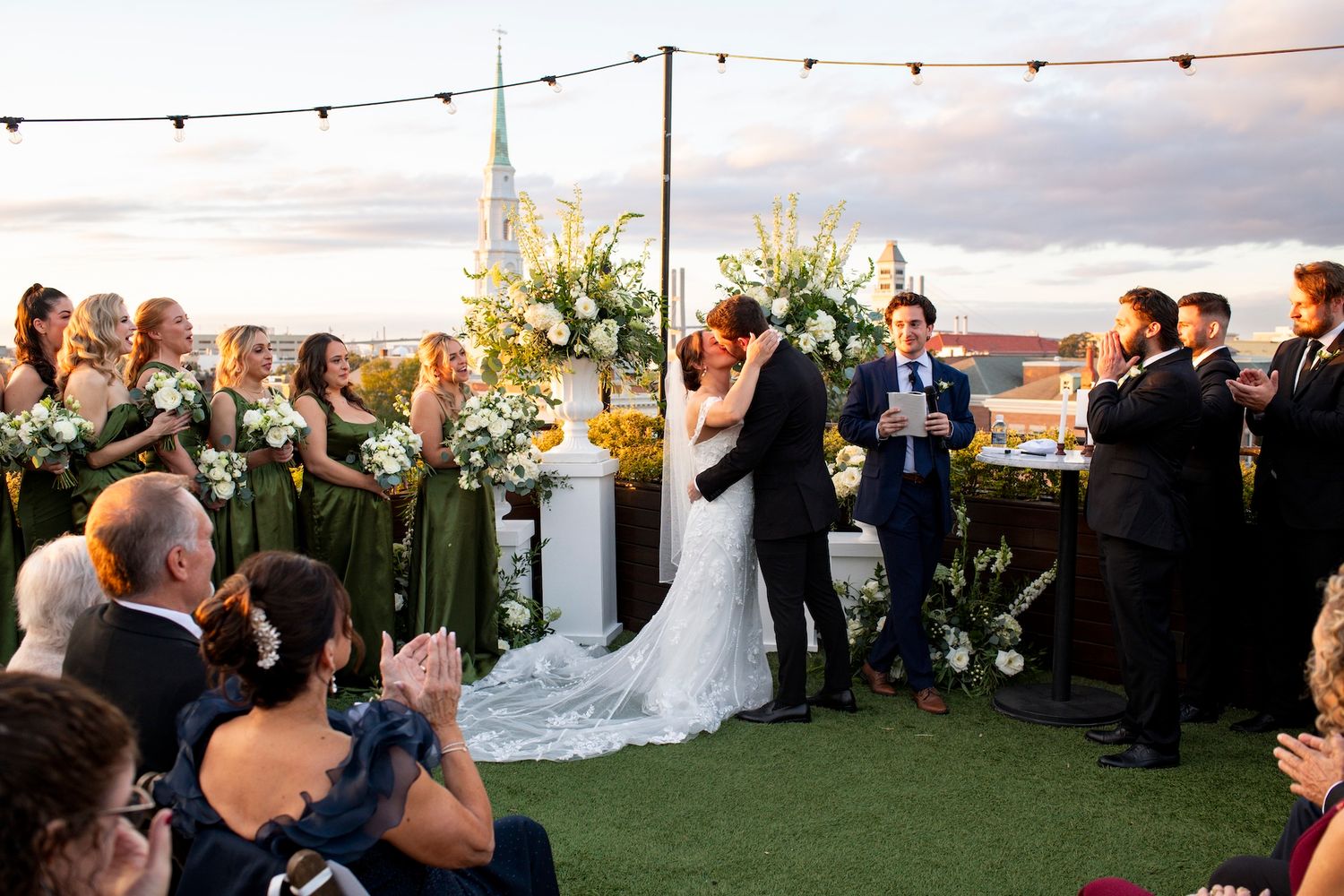 String lights illuminate an outdoor evening wedding ceremony with guests seated on a grassy lawn at sunset.