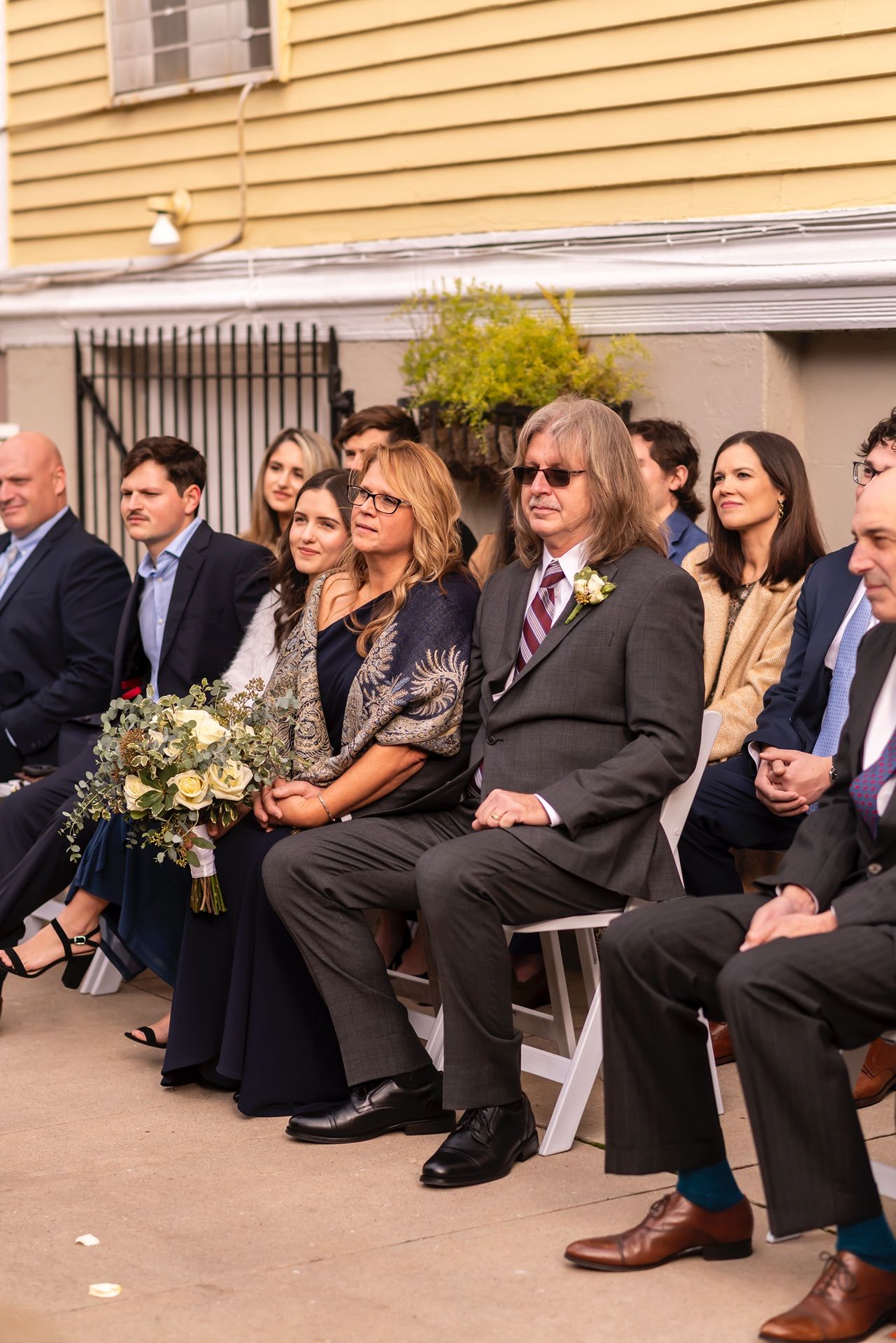 A group of guests sit together in formal attire at an outdoor event with a yellow building in the background.